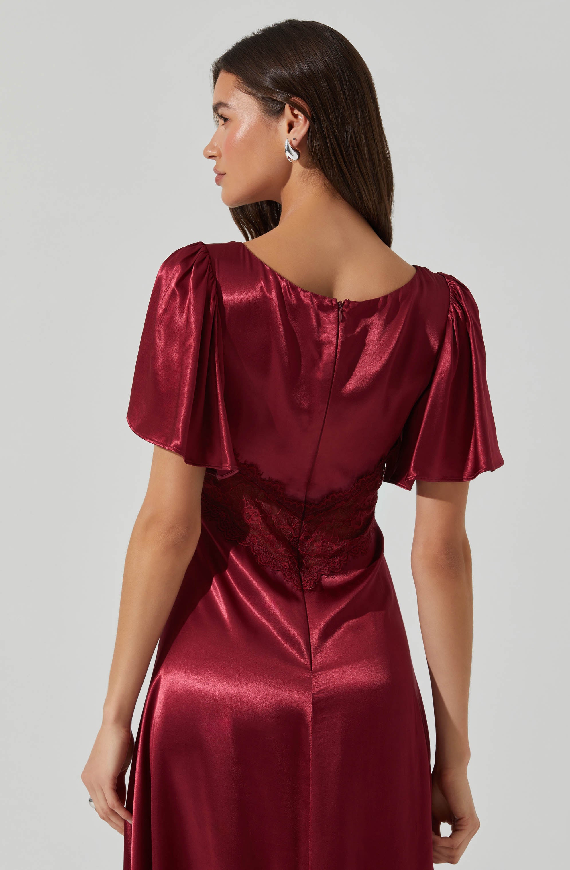 A woman is wearing a deep red satin dress with puffed short sleeves that have gathered fabric at the shoulders. The dress features a fitted waist with lace detailing and a zipper running down the back. The fabric has a shiny, smooth finish.