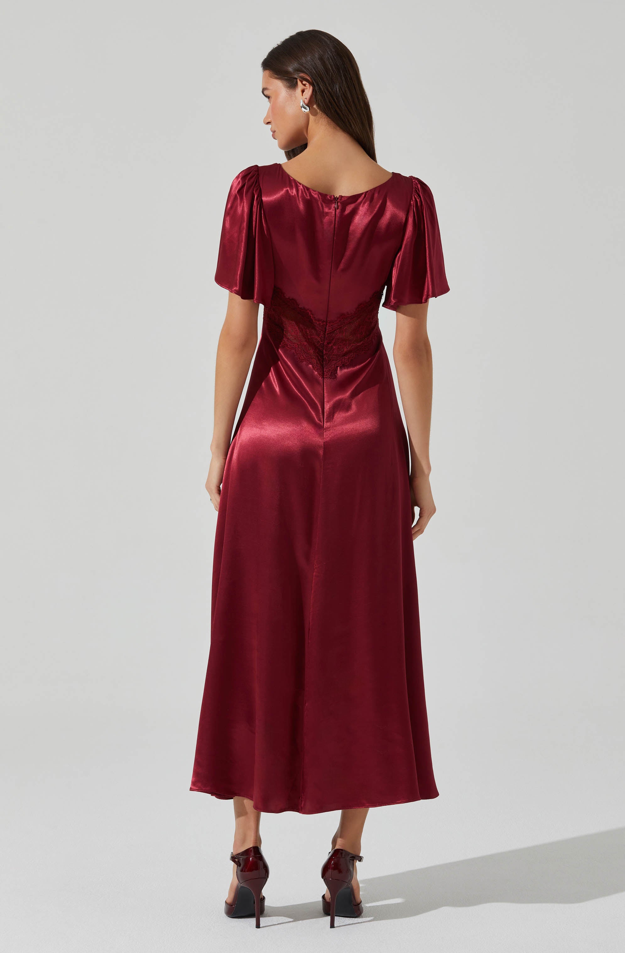 A woman is wearing a deep red satin dress with a fitted bodice and wide-leg skirt. The dress features puffed short sleeves with gathers at the shoulder seam. The back of the dress has a zipper closure, and the waist is accented with a lace inset. She is also wearing matching high-heeled shoes.