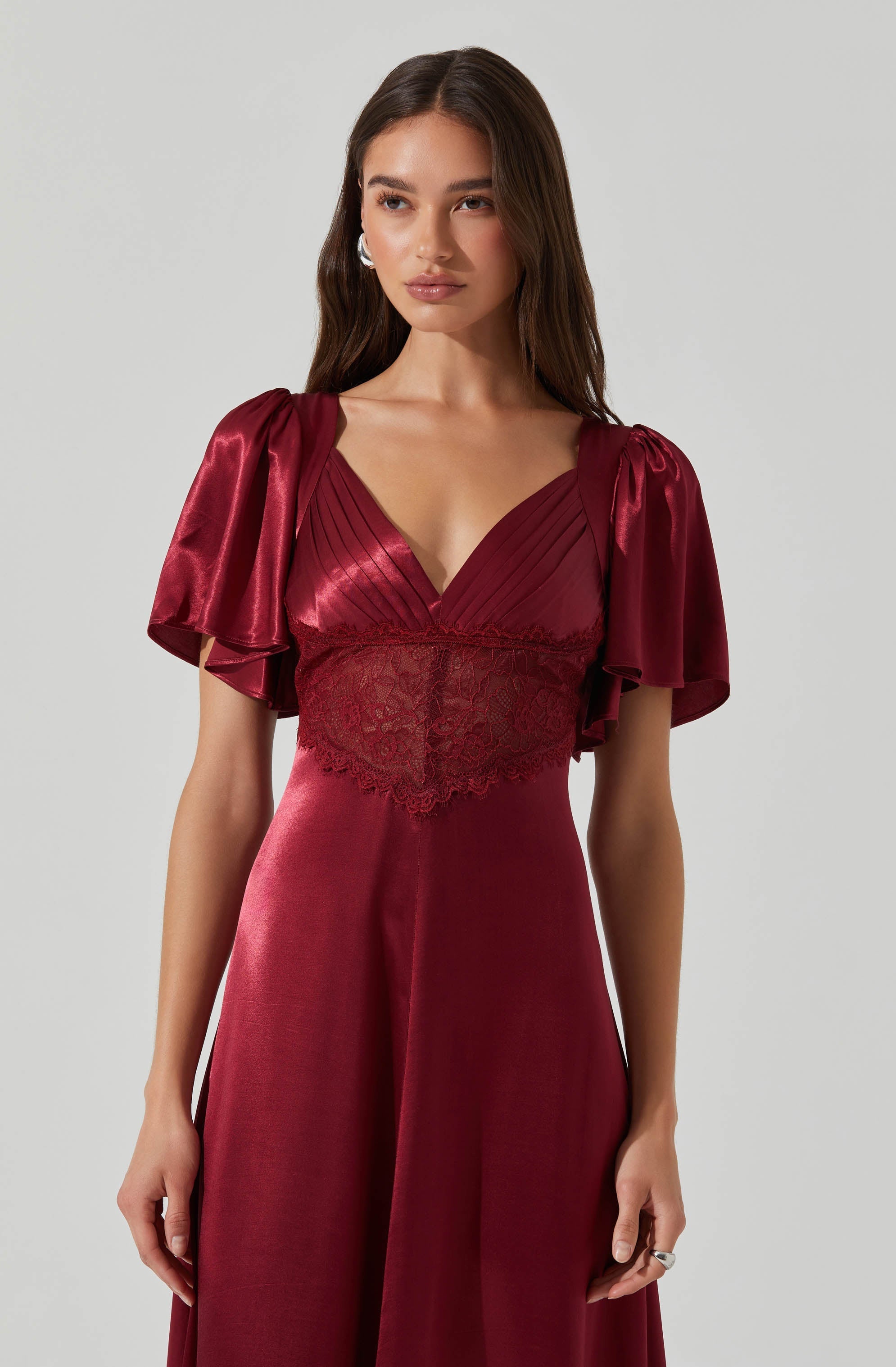 The dress is a deep red, satin-like fabric with a V-neckline. It features puffed, short sleeves made from shiny, smooth fabric that drape loosely. The bodice has vertical pleats and a lace insert at the waist area, adding texture and detail. The skirt is long, flowing, and made from the same satin-like material, creating an elegant silhouette.