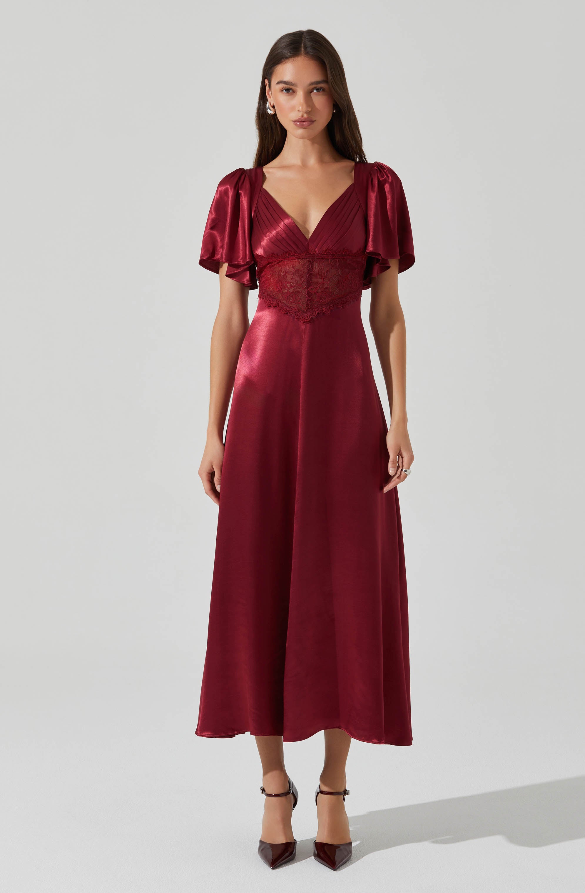 A woman is wearing a deep red satin evening gown with short, puffed sleeves made of a shiny, textured fabric. The dress features a V-neckline and a fitted bodice with lace details, flowing into a long, wide skirt that reaches her ankles. She is also wearing pointed-toe heels with ankle straps.