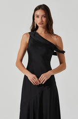 A woman is wearing a sleek, black, one-shoulder dress made of shiny satin fabric. The dress features a draped detail on the shoulder and fits snugly at the waist, flowing down into a long skirt. She is accessorized with long, dangling earrings and a ring on her right hand, with her long, wavy brown hair parted on the side and resting over her shoulders.