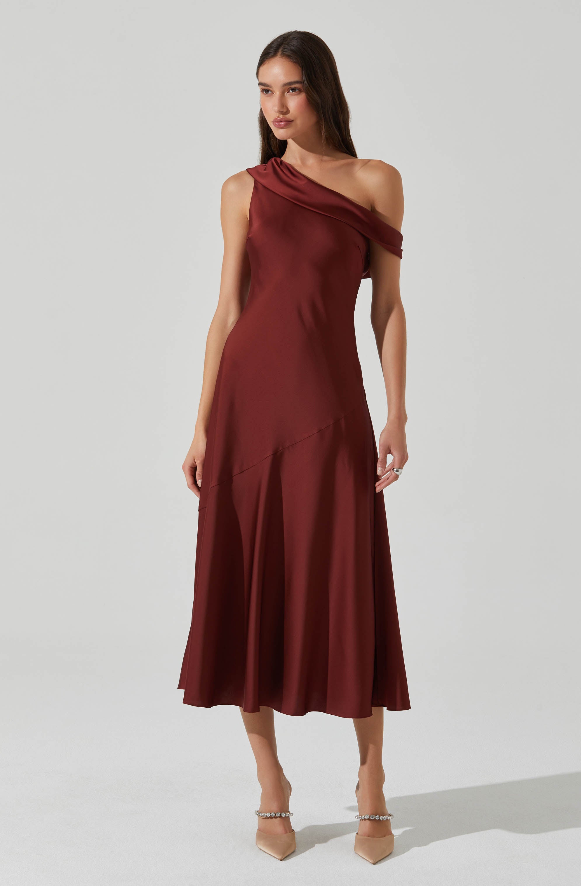 A woman is wearing a sleeveless, burgundy satin dress with an asymmetrical, draped neckline that extends over one shoulder. The dress has a fitted waist and flows down to a midi length with a subtle diagonal seam detail. She is also wearing nude high-heeled shoes with pearl embellishments around the ankle.