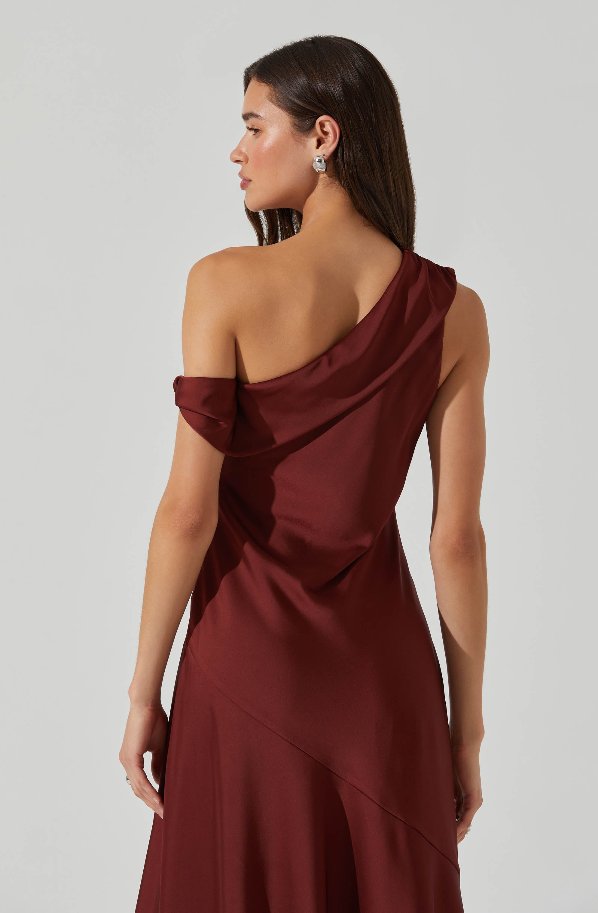 A woman is wearing an elegant, floor-length gown made of shiny satin fabric in a deep burgundy color. The gown features an asymmetrical, one-shoulder design with a draped, off-the-shoulder sleeve on her left arm, while her right shoulder is bare. The fabric appears smooth and glossy, adding a sophisticated touch to the overall look. She accessorizes with large, silver statement earrings.