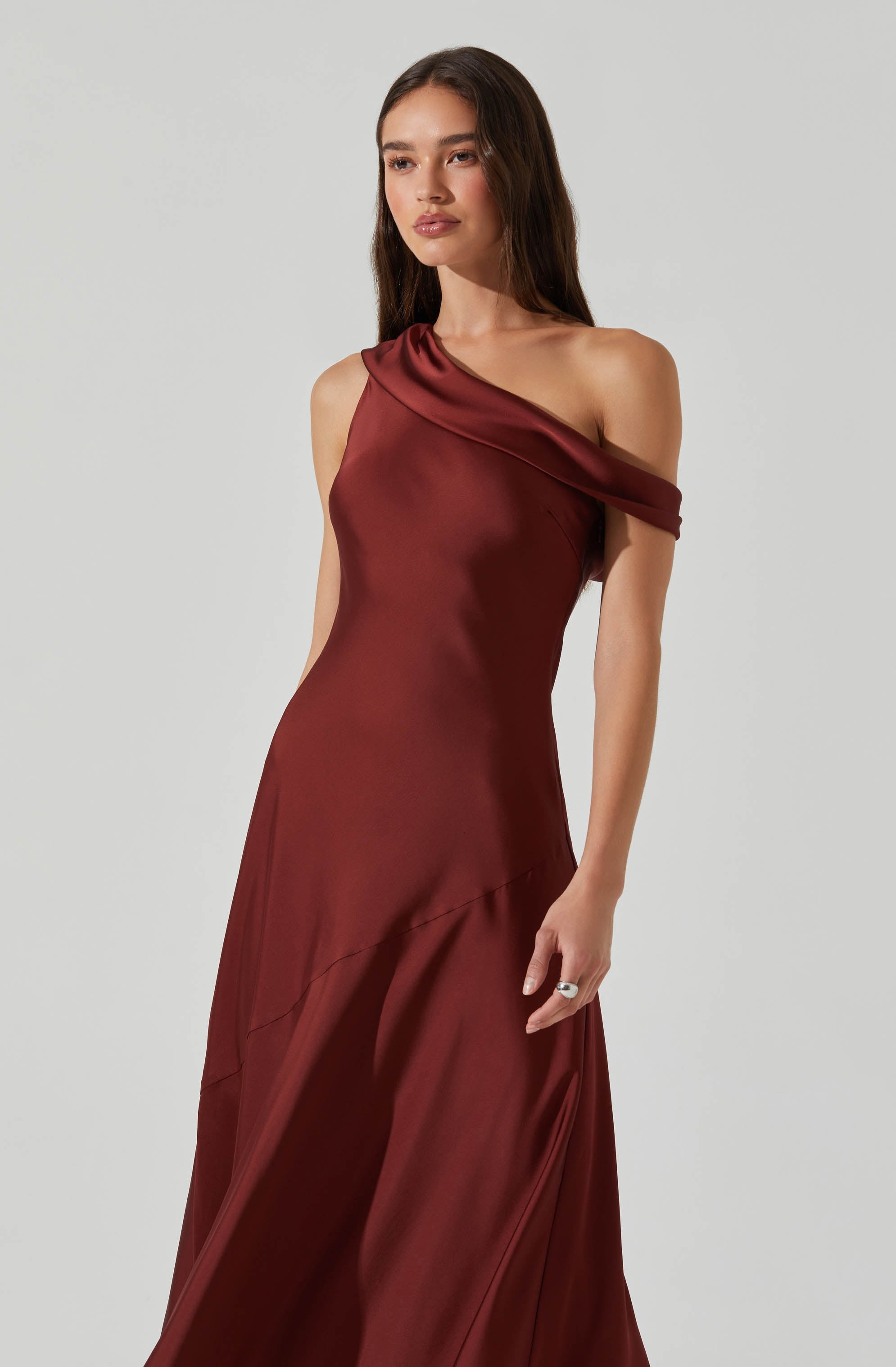 A woman is wearing a sleeveless, floor-length dress made of shiny, satin-like fabric in a deep burgundy color. The dress features an off-shoulder design with a draped detail over one shoulder.