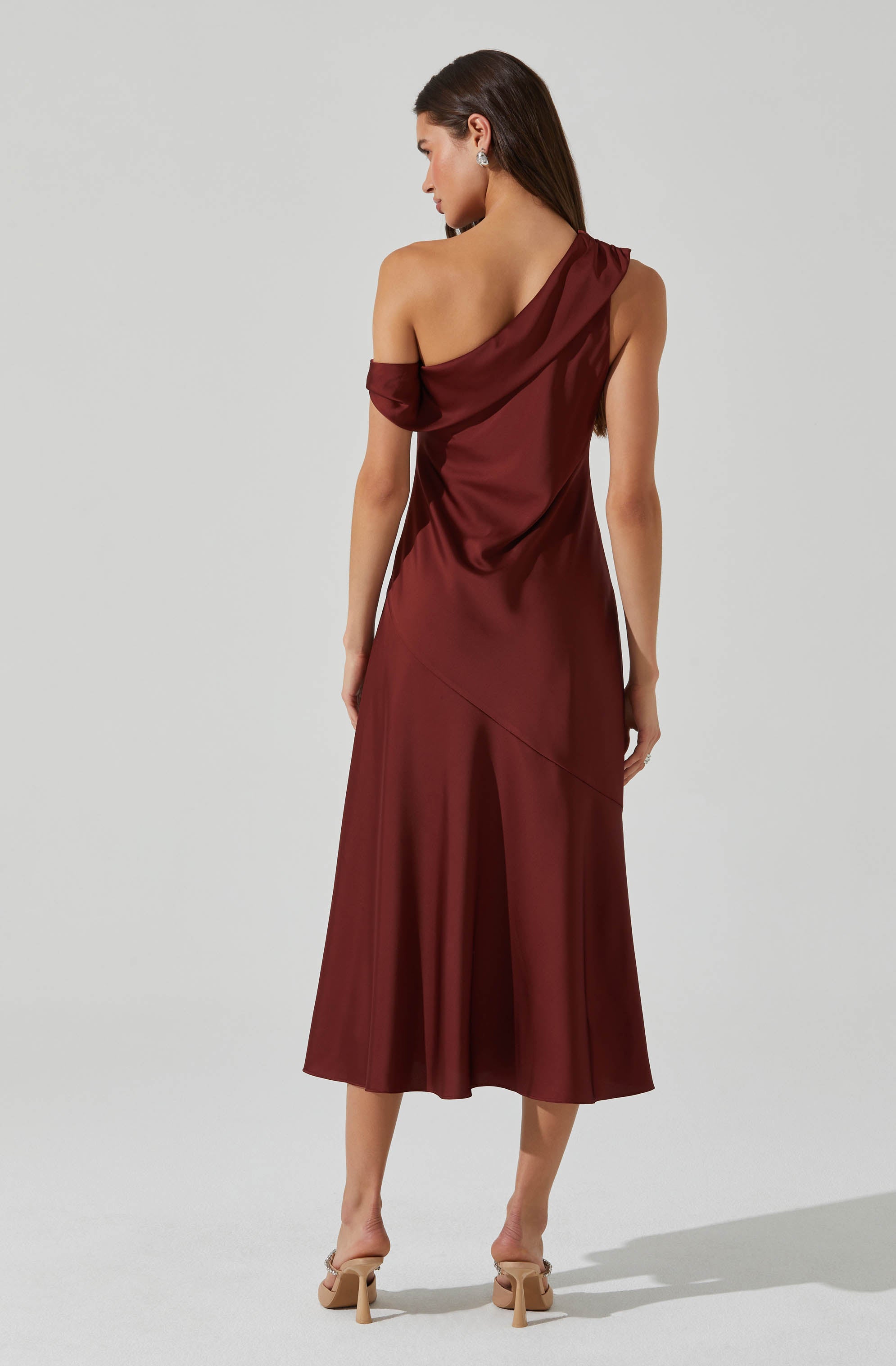 A woman is wearing a sleeveless, deep burgundy satin dress with an asymmetrical, draped neckline. The dress has a fitted bodice and flows into a midi-length skirt with a slight flare and an angled seam detail across the hip. She pairs the dress with beige high-heeled shoes.