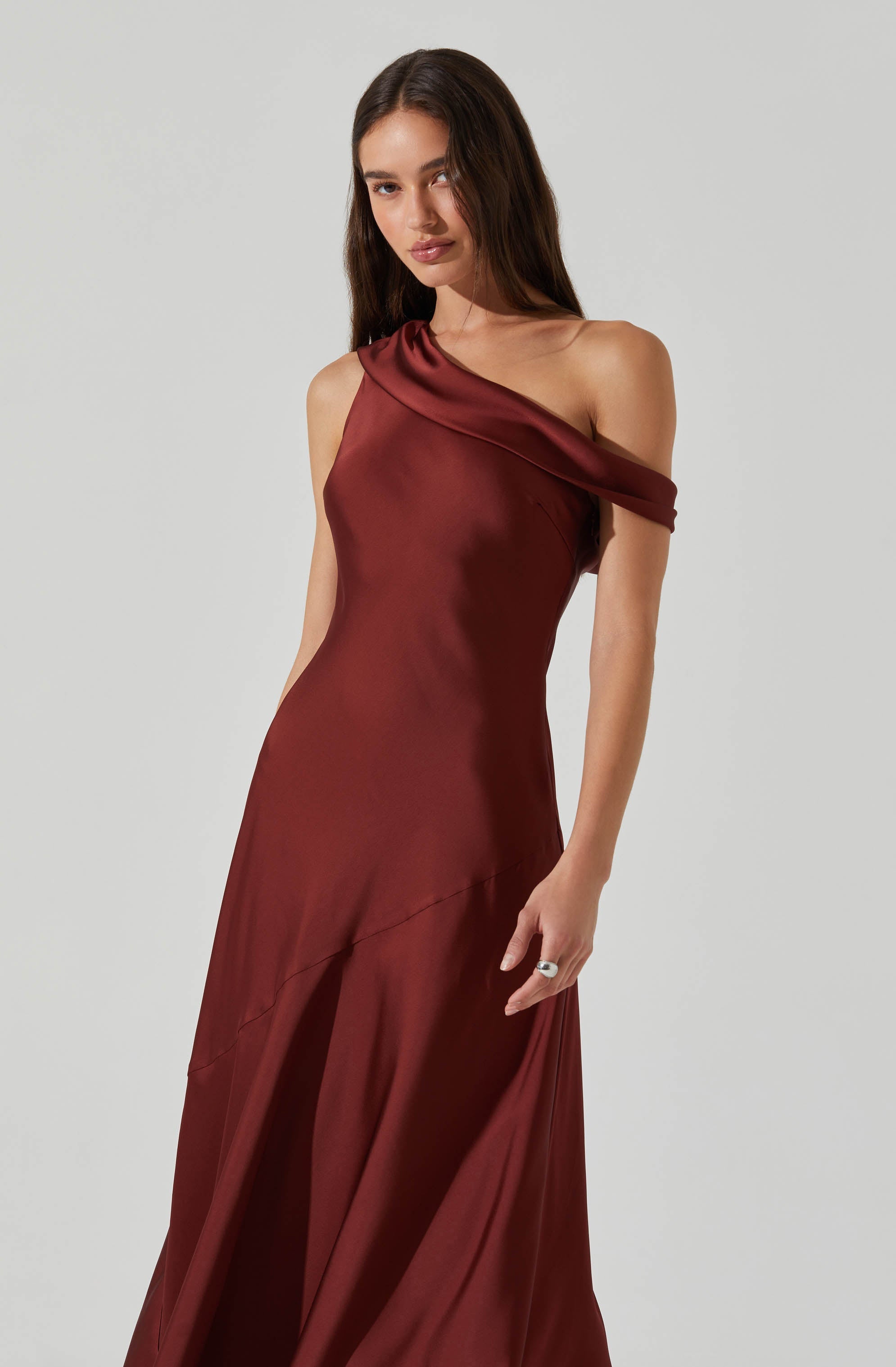 A woman is wearing a floor-length, burgundy satin gown with a one-shoulder design. The dress features a draped neckline that extends over her shoulder, creating an elegant, asymmetrical look. The fabric has a smooth, shiny finish and the gown appears fitted through the waist, flowing down gracefully.