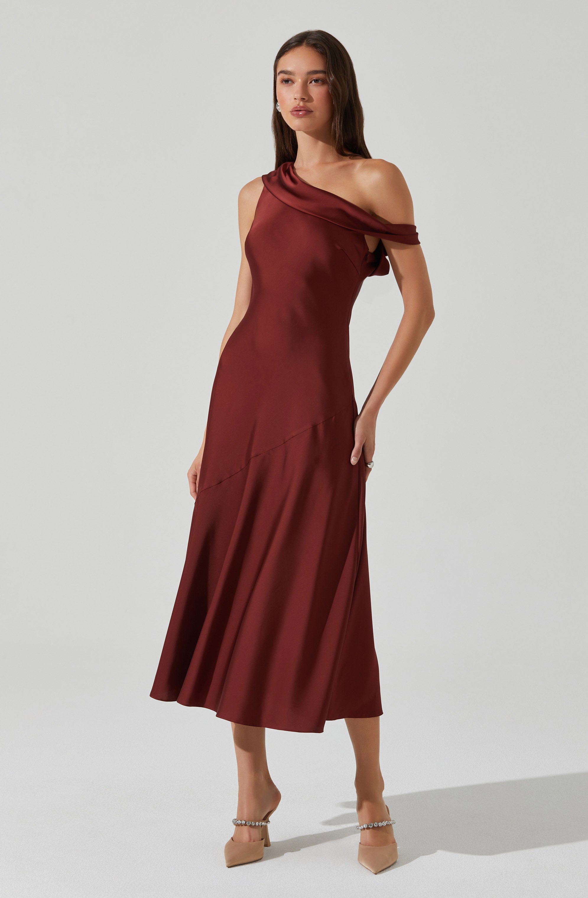 A woman is wearing a floor-length, satin burgundy dress with an asymmetrical design. The dress features a one-shoulder neckline with a draped fabric detail across the shoulder, creating an off-the-shoulder look on one side. The fabric appears smooth and shiny, with a subtle diagonal seam detail running across the skirt. She pairs the dress with nude-colored heels that have pearl embellishments.