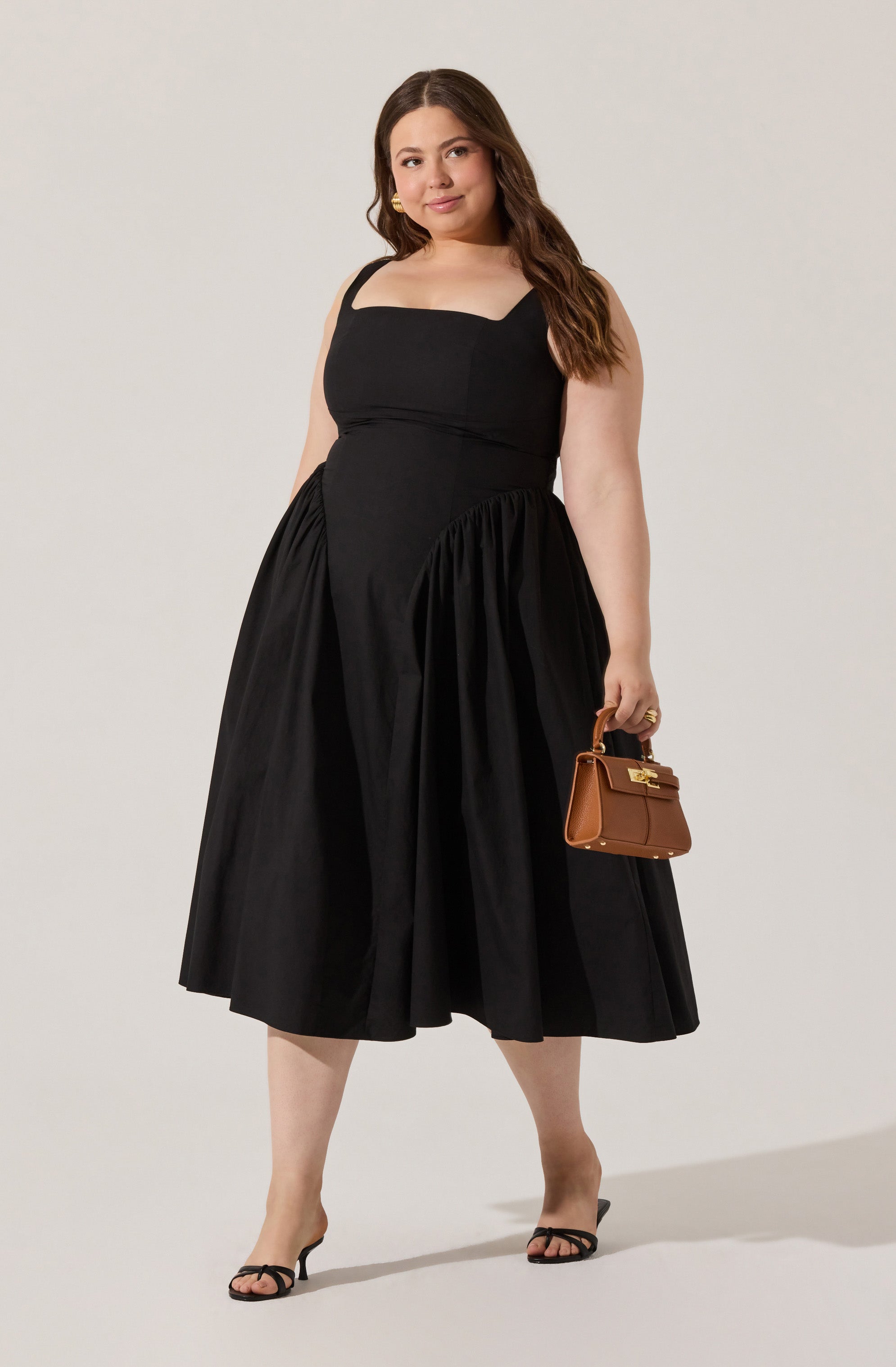 A woman is wearing a sleeveless, black midi dress with square neckline and wide straps. She is holding a small, brown handbag with a top handle and a gold clasp. She is also wearing black open-toe high-heeled sandals.