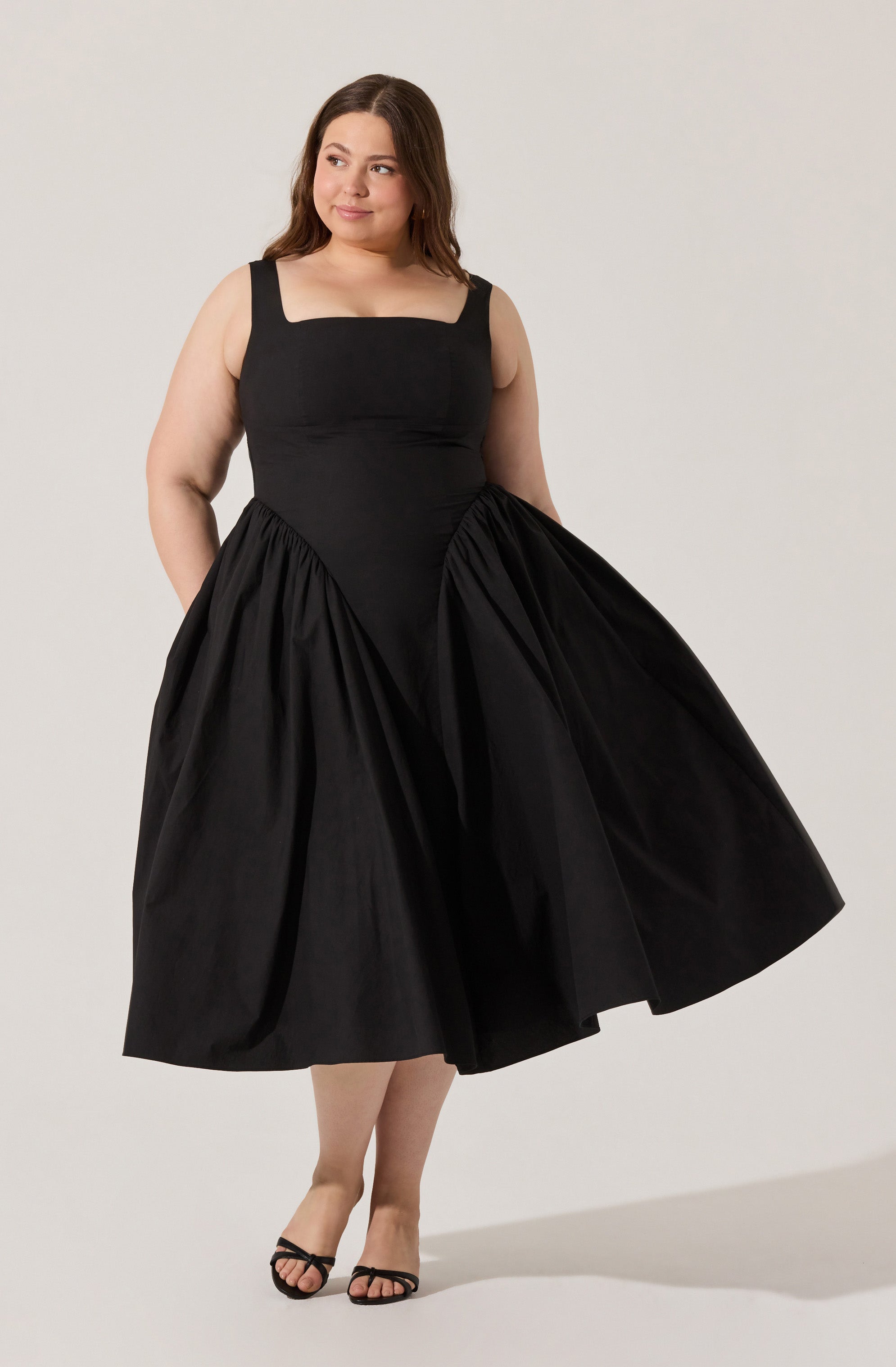 A woman is wearing a sleeveless black dress with a fitted bodice and wide straps. The dress features a full, voluminous skirt that reaches mid-calf length. She is also wearing black open-toe high-heeled sandals.