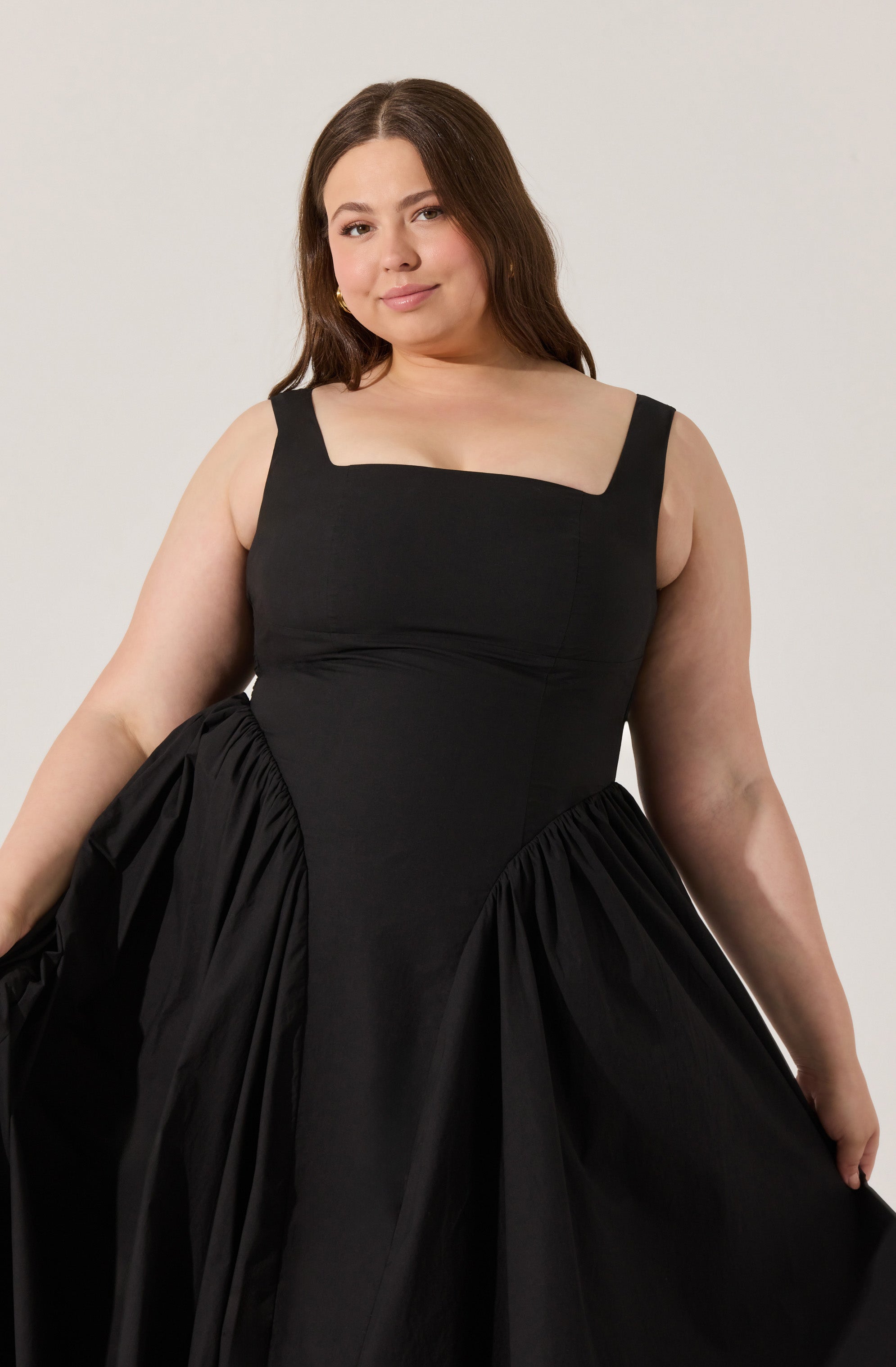 A woman is wearing a black dress with wide straps and a square neckline. The dress has a fitted bodice and a full, gathered skirt that creates a voluminous silhouette.