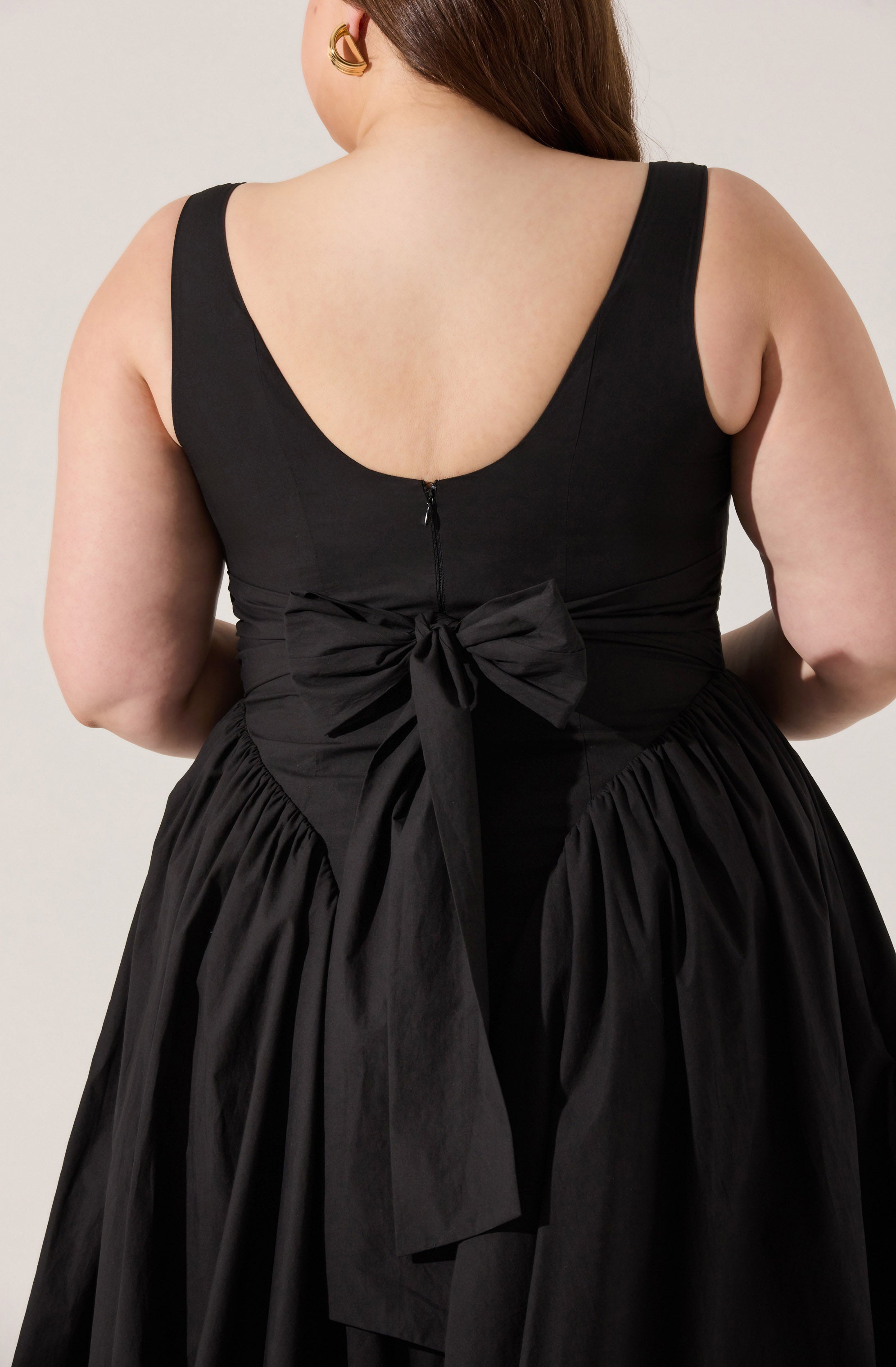 A woman is wearing a sleeveless, fitted black dress with a scoop back neckline. The dress features a large bow tied at the waist in the back, and the skirt is gathered with pleats, creating a full, voluminous look. She has gold hoop earrings as accessories.