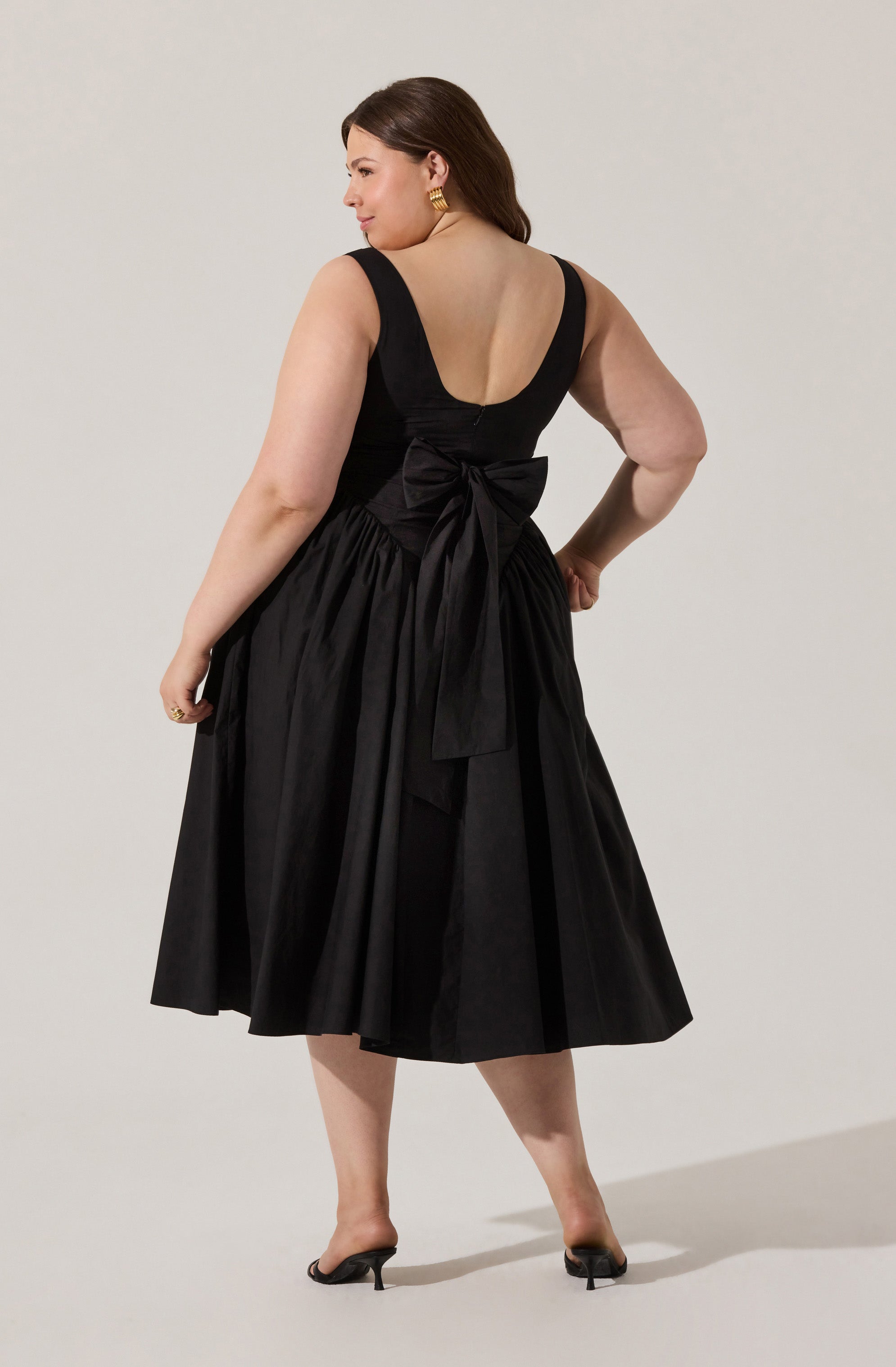 A woman is wearing a sleeveless black dress with a deep scoop back. The dress has a fitted bodice and a full, gathered midi skirt. There is a large bow tied at the waist, made from the same fabric as the dress. She is also wearing gold earrings and black open-toe heels.
