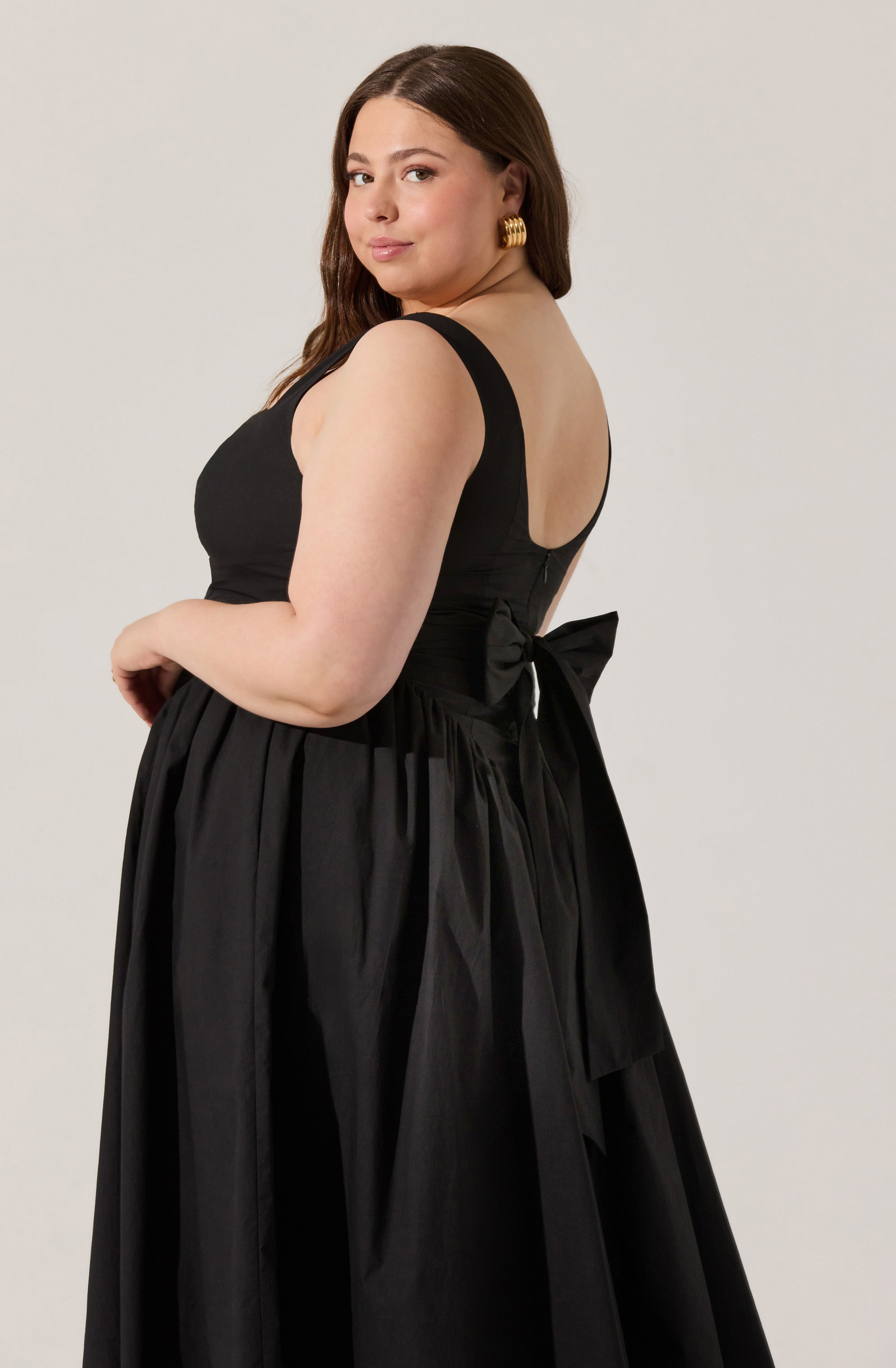 A woman is wearing a sleeveless, fitted black top with broad shoulder straps, paired with a high-waisted, full-length black skirt that has gathered fabric at the waist and is tied with a large bow at the back. She has gold hoop earrings and her long brown hair is styled in loose waves.