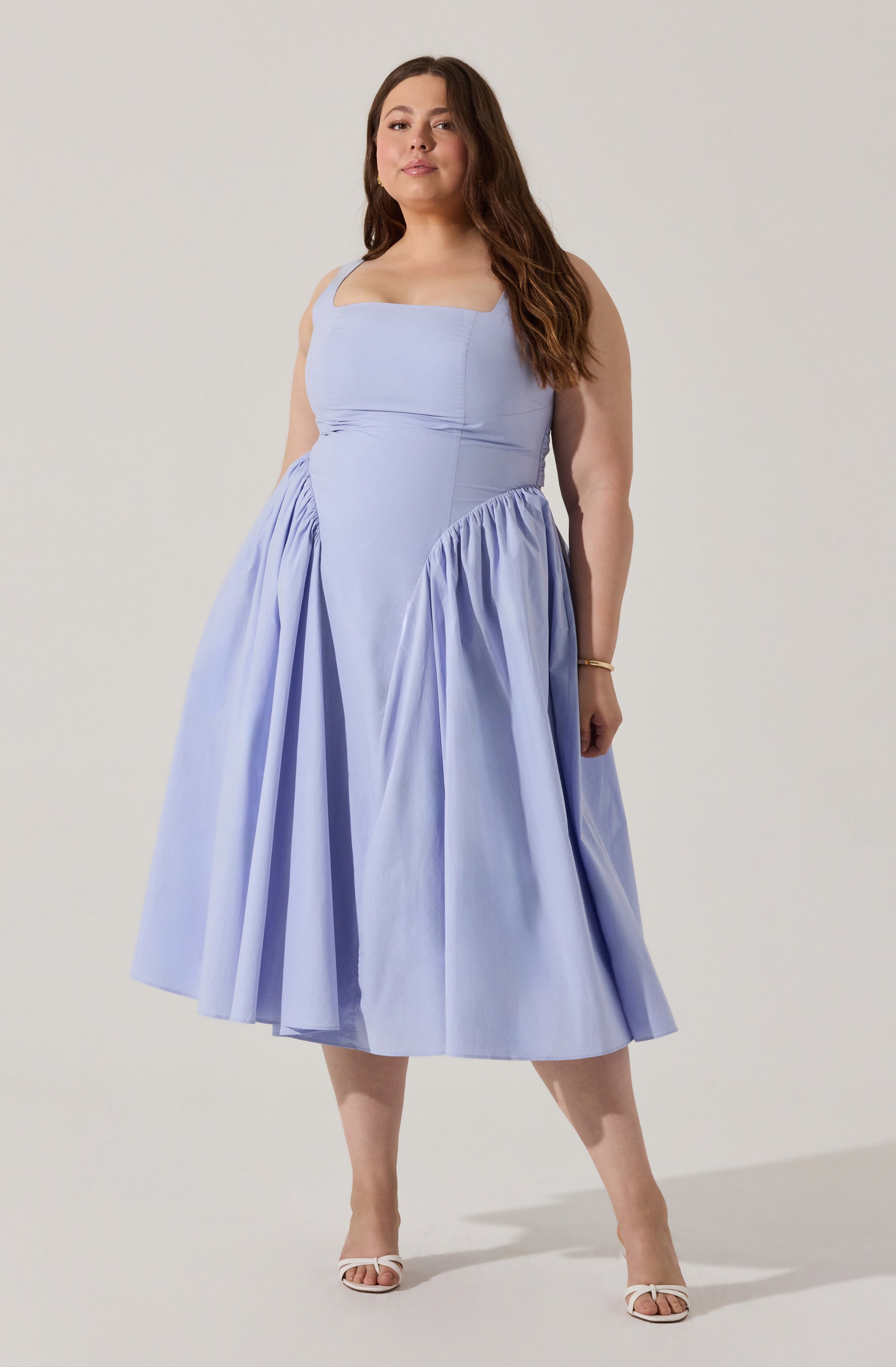 A woman is wearing a sleeveless, light blue midi dress with a fitted bodice and wide straps. The dress features gathered fabric around the waist, creating a full, pleated skirt that falls below the knees. She is also wearing white open-toe heels and a gold bracelet on her right wrist.