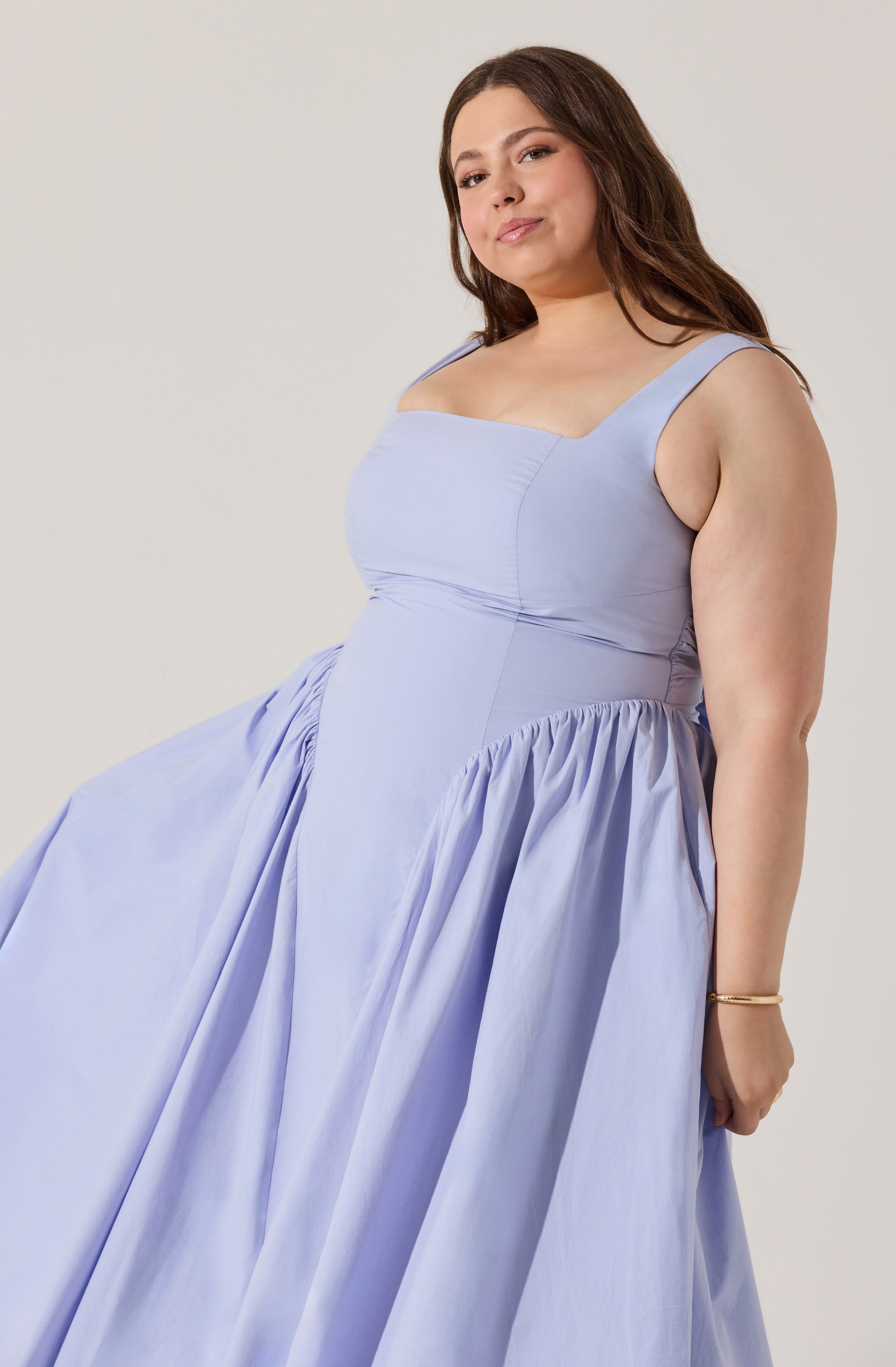 A woman is wearing a sleeveless, light lavender dress with a fitted bodice and wide shoulder straps. The dress has a gathered, full skirt that extends to the floor. The fabric appears smooth and slightly shiny.