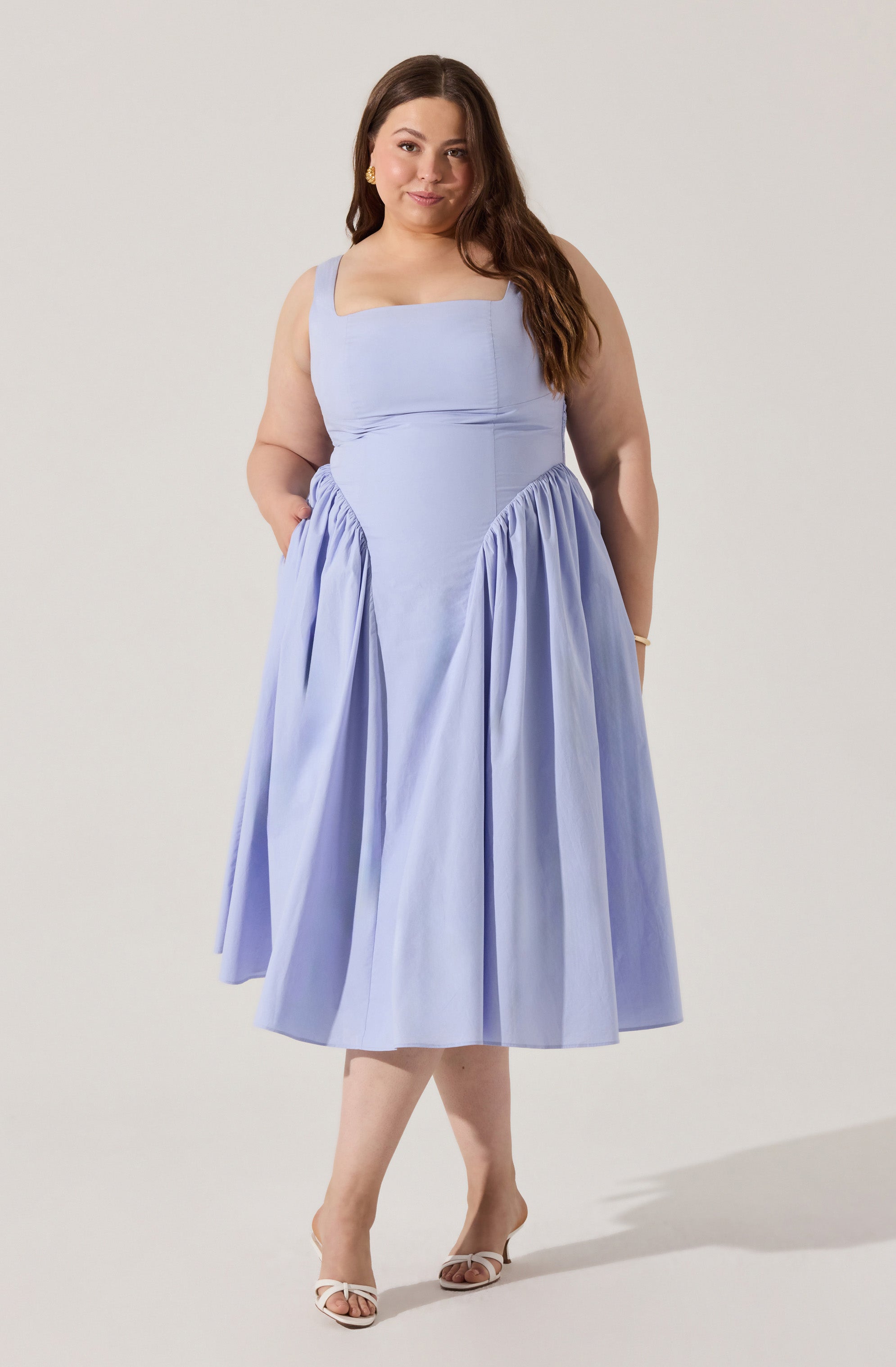 A woman is wearing a sleeveless, light lavender dress with a fitted bodice and wide shoulder straps. The dress features a gathered, full skirt that falls below the knees. She is also wearing open-toe, high-heeled shoes with a bow detail on the front.