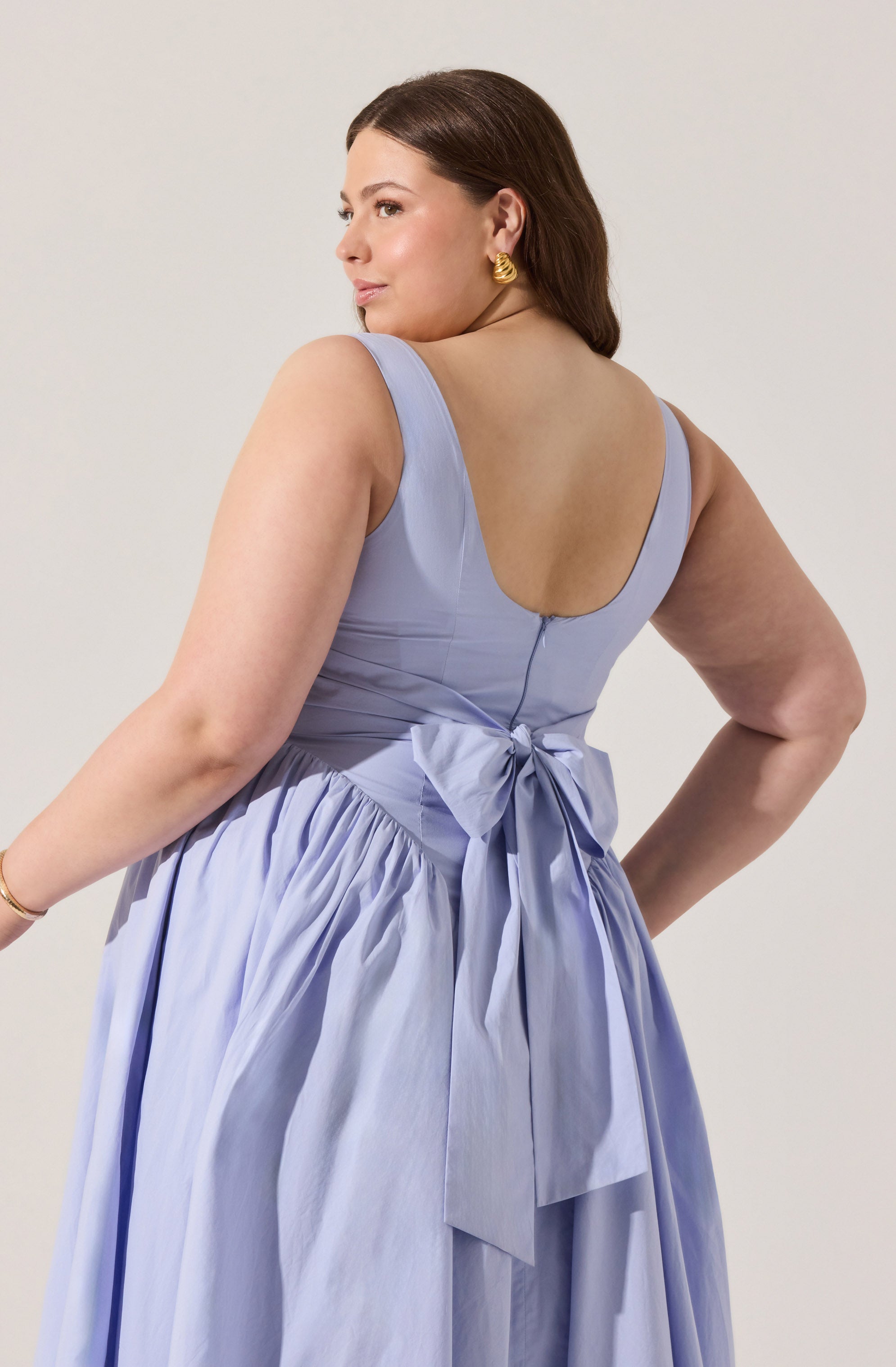 A woman is wearing a sleeveless, lavender-colored dress with a fitted bodice and a scoop neckline. The dress features a large bow tied at the back waist, with the fabric gathered into pleats that extend down the skirt. She has dark brown hair styled in soft waves and is accessorized with gold hoop earrings and a gold bracelet.