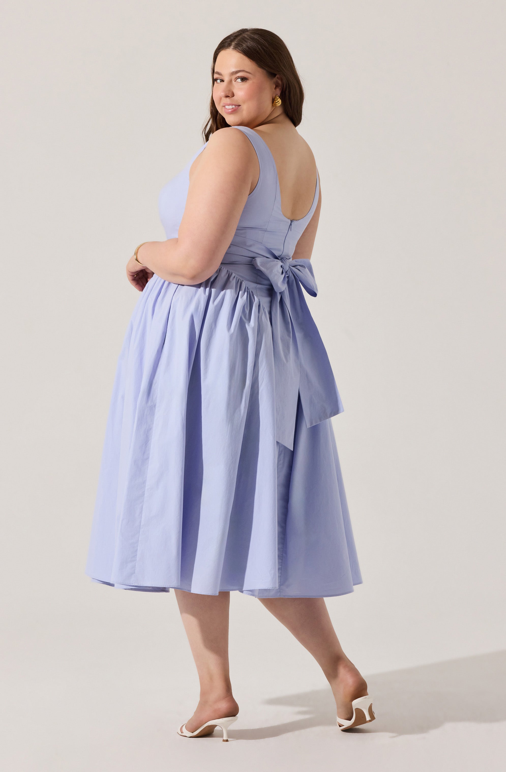 A woman is wearing a sleeveless, light blue dress with a fitted bodice and a full, gathered skirt that falls below the knees. The back of the dress features a large bow at the waist and a zipper closure. She is also wearing a pair of white high-heeled sandals with open toes and a gold bracelet. She has dark, wavy hair and is accessorized with gold earrings.