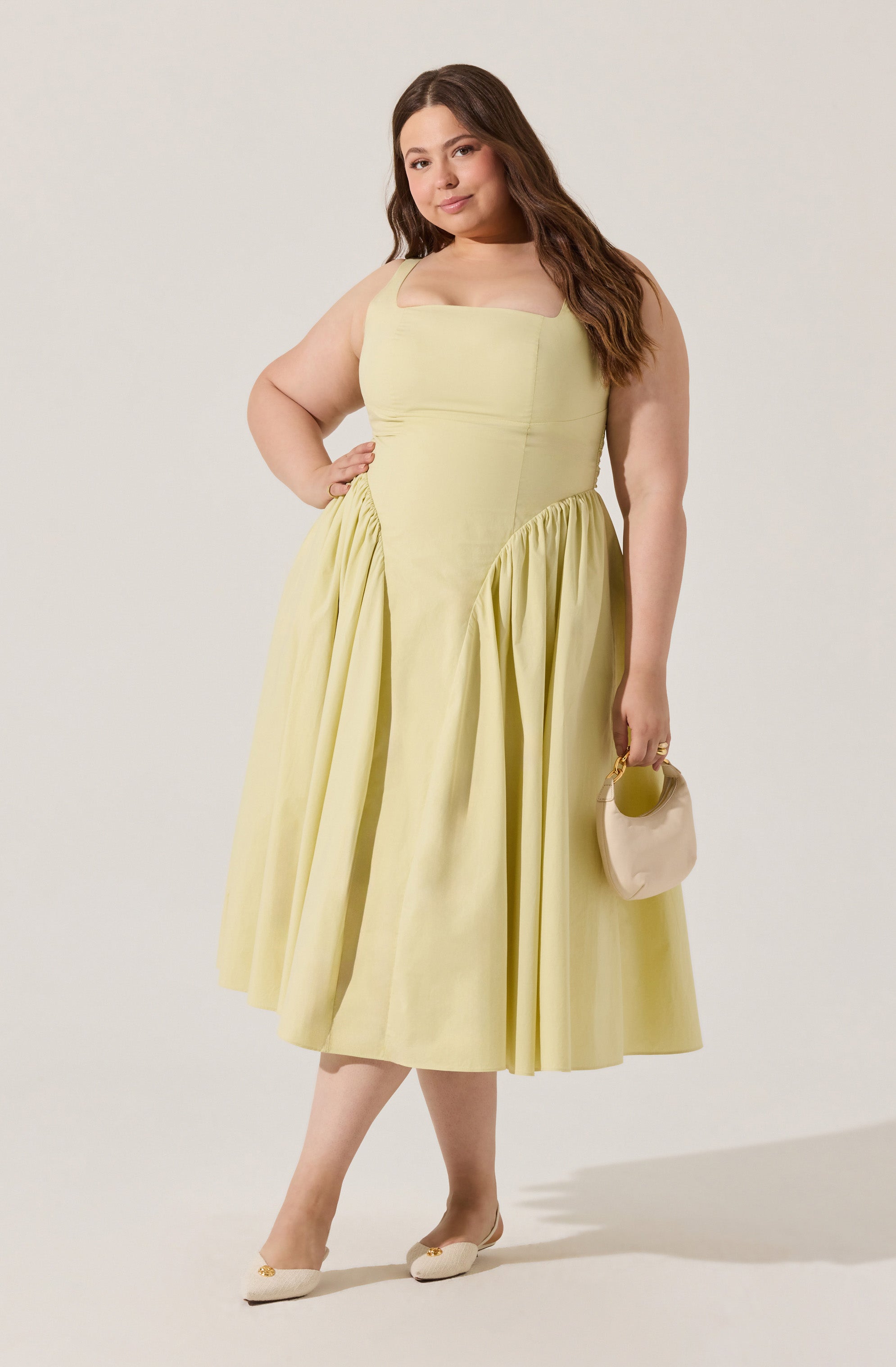 A woman is wearing a sleeveless, pale yellow, midi-length dress with a fitted bodice and a full, gathered skirt. She holds a small, beige handbag with a circular handle in her right hand, and she is wearing beige shoes with a small heel and decorative embellishments.