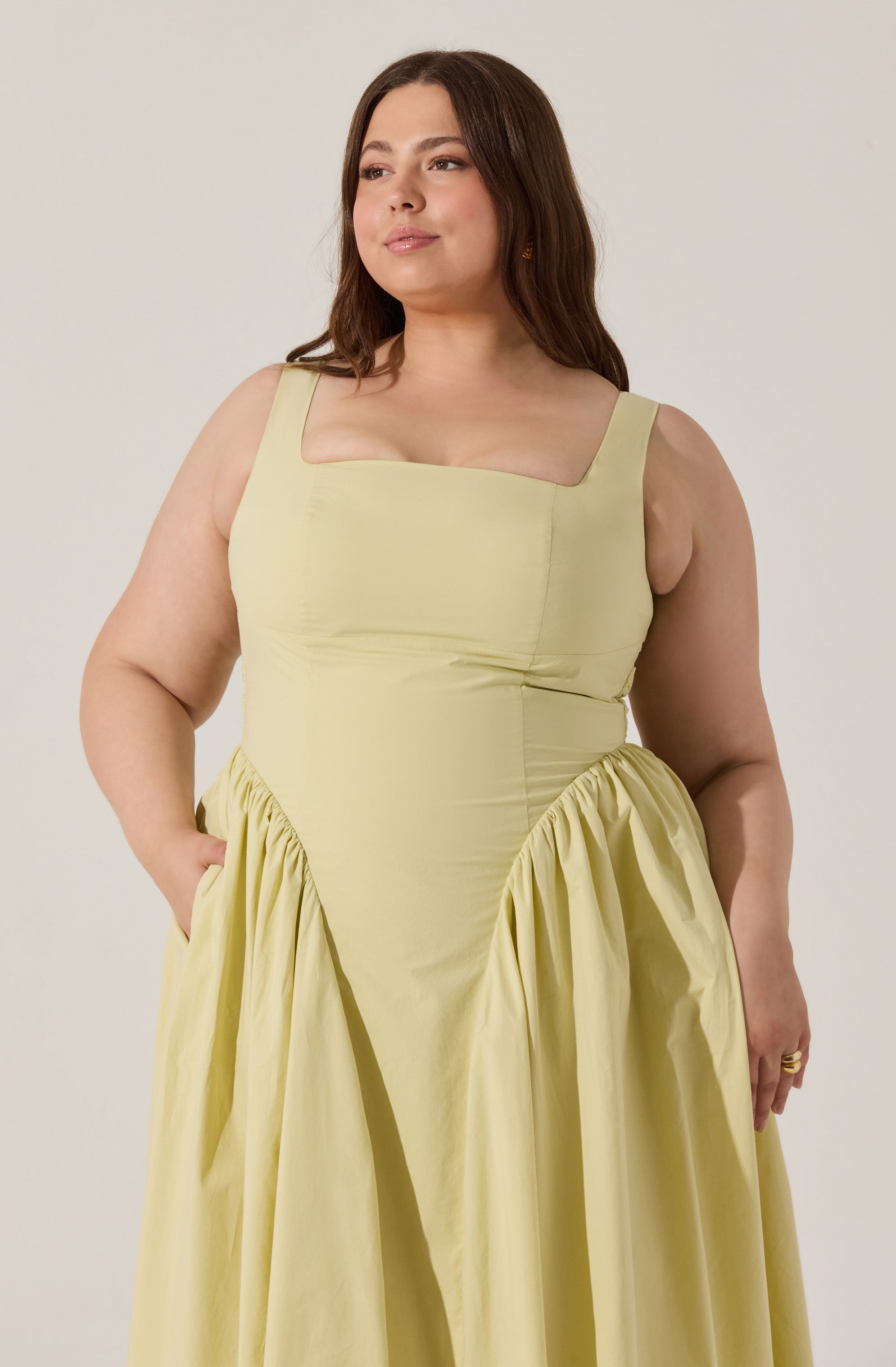 A woman wearing a sleeveless, pale yellow dress with a fitted bodice and a square neckline. The dress features gathered fabric details on the sides of the skirt, creating a voluminous, draped effect. The shoulder straps are wide and the dress appears to be made of a smooth, lightweight fabric.
