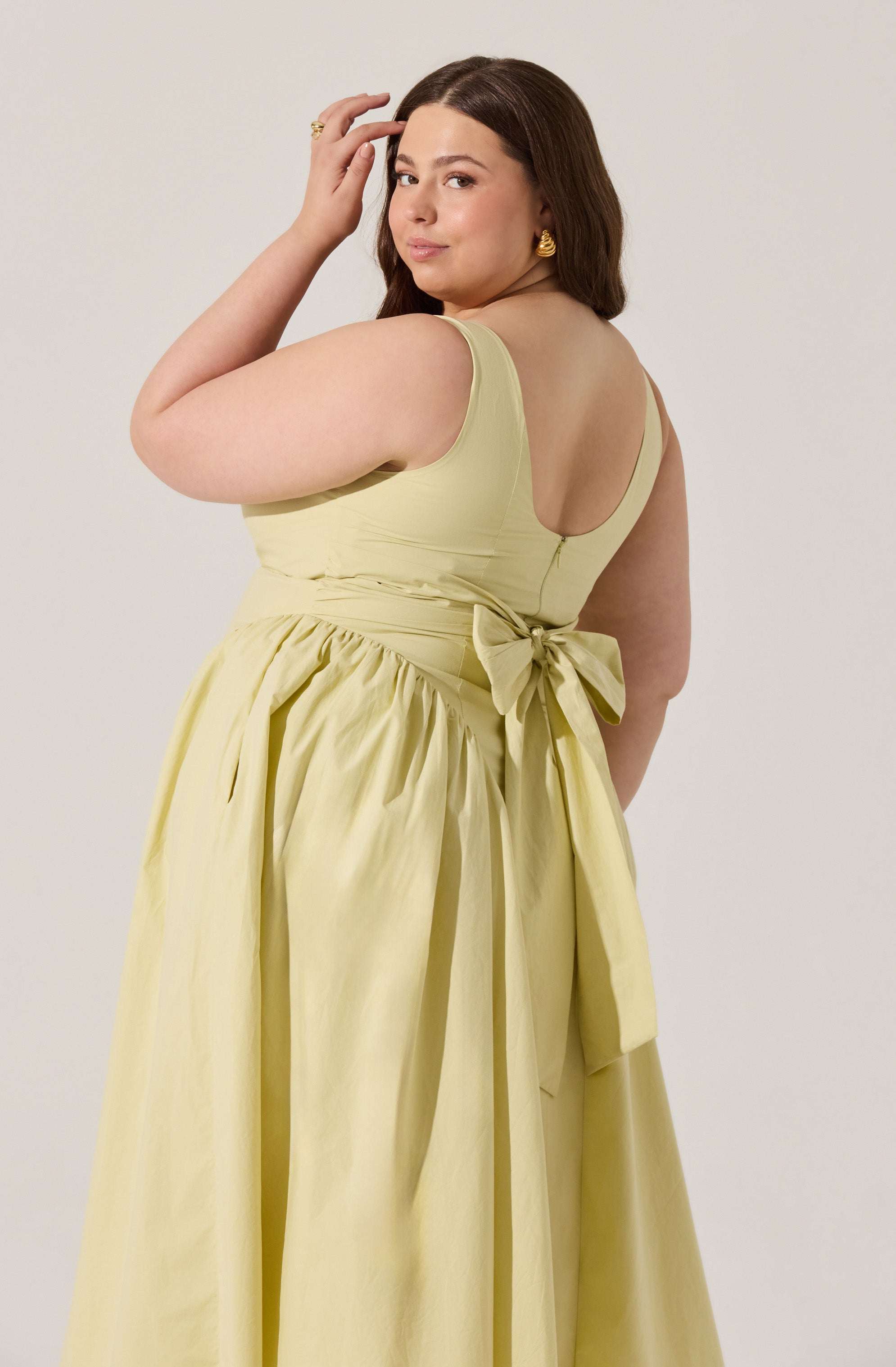 The dress is sleeveless with a fitted bodice and a scoop neckline. It is a light yellow color with a wide sash around the waist that ties into a bow at the back. The skirt is full and gathered, creating a voluminous look.