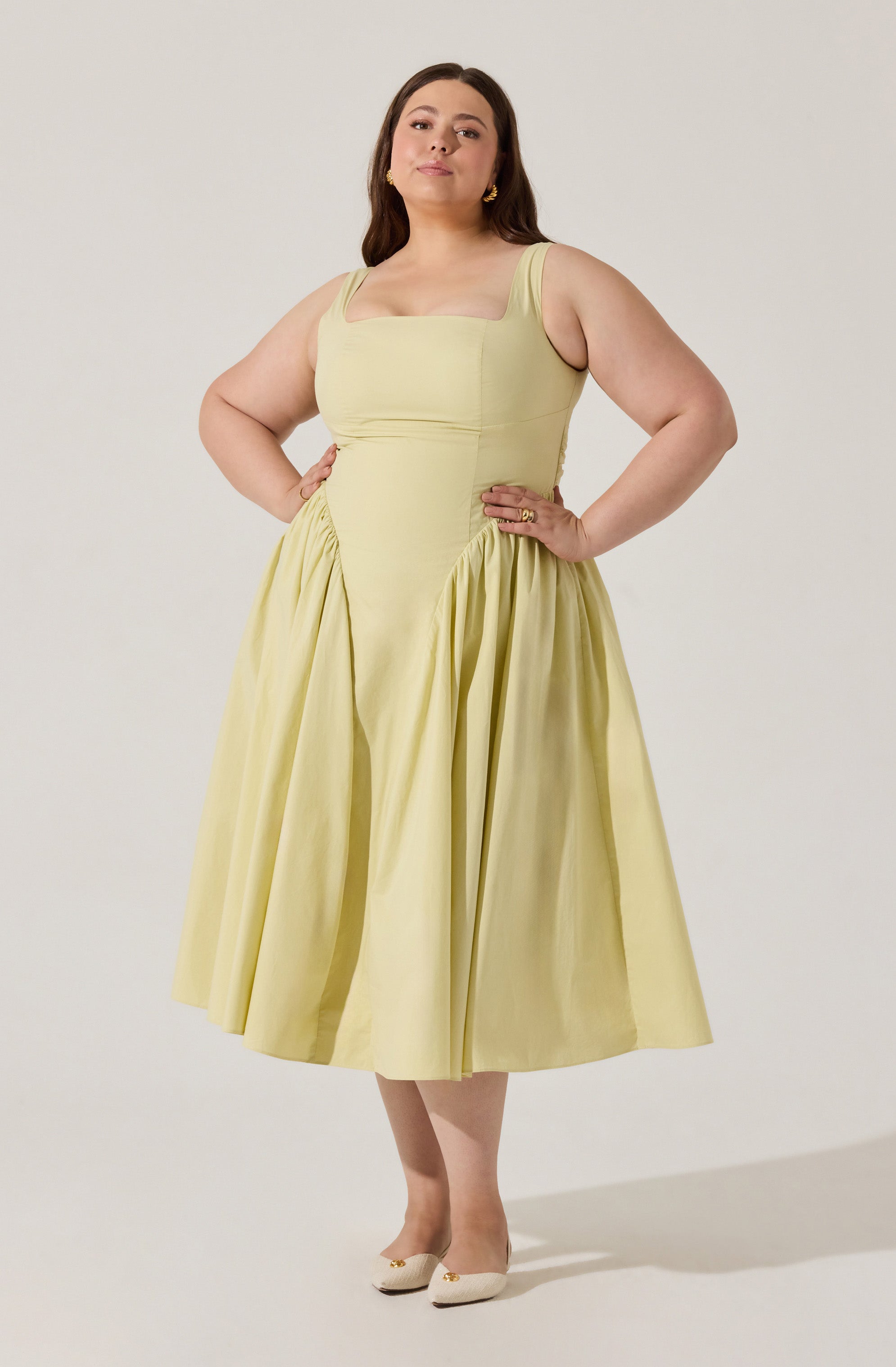 A woman is wearing a sleeveless, light yellow dress with wide shoulder straps. The dress has a fitted bodice with a square neckline and a full, gathered skirt that falls below the knees. She pairs the dress with beige shoes that have a slight heel and decorative details on the front.