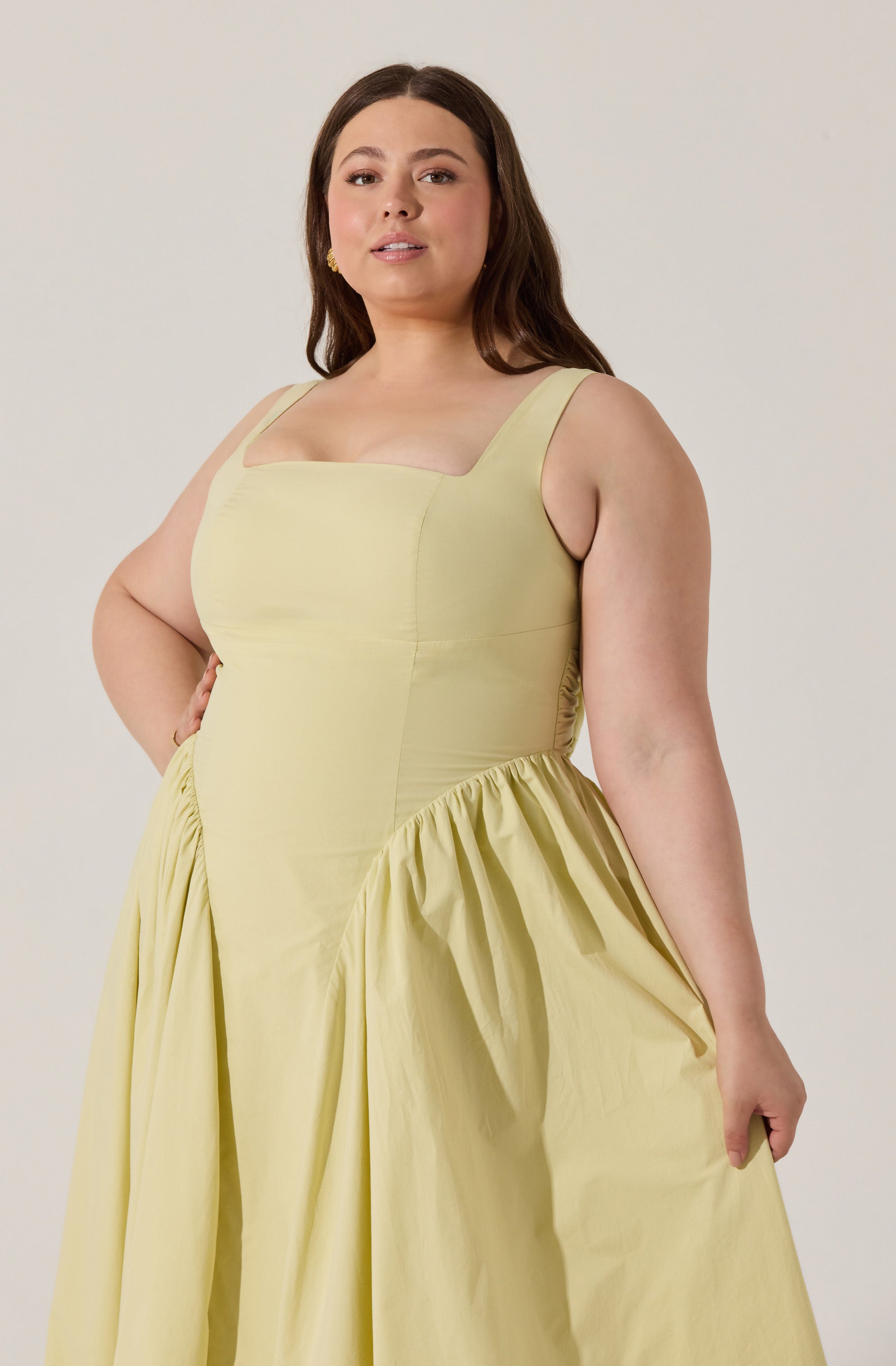 A woman wearing a sleeveless, light yellow dress with wide straps. The bodice is fitted with a square neckline, and the skirt features gathered fabric creating volume and draping.