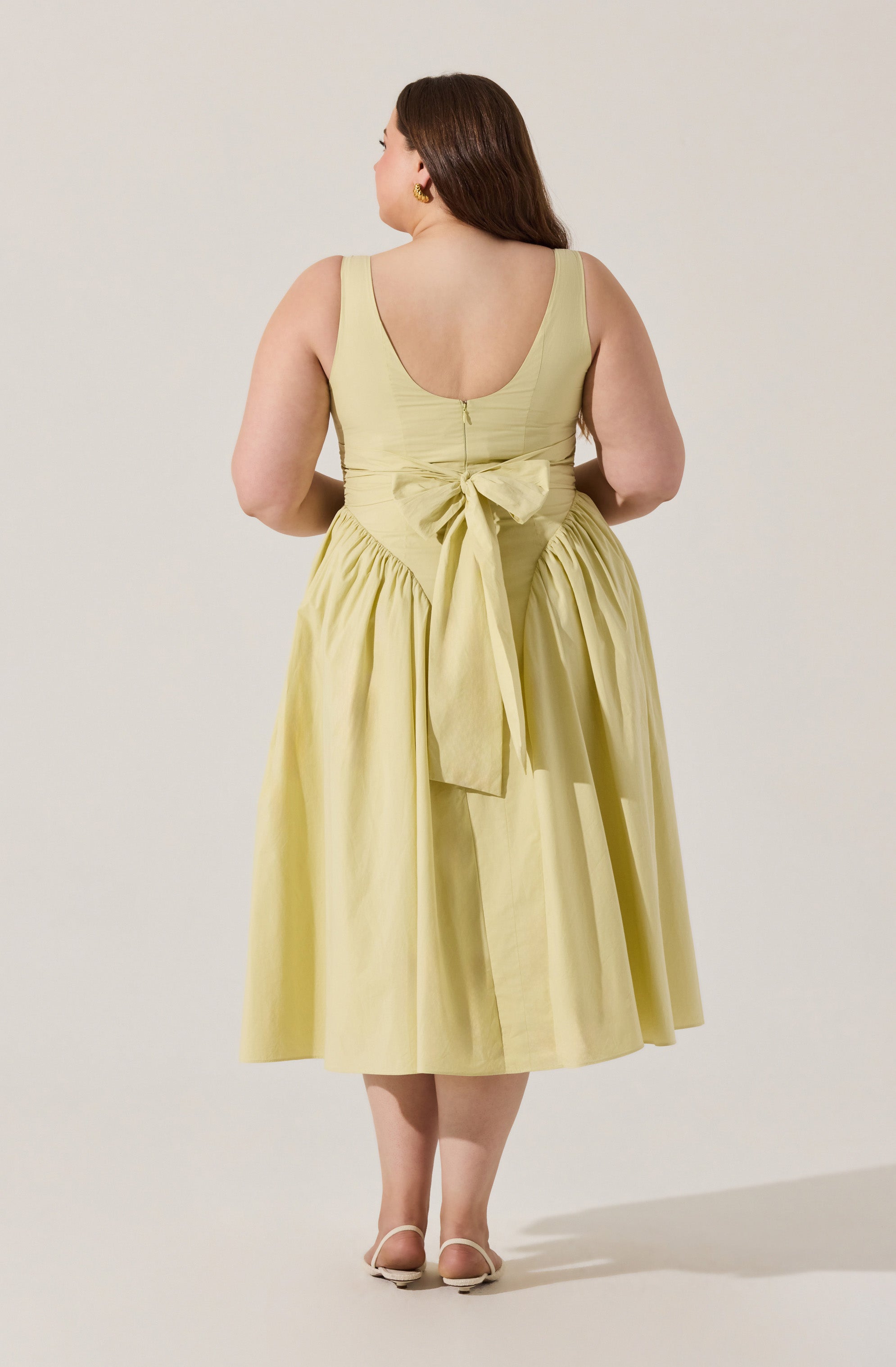 A woman is wearing a sleeveless, light yellow dress with a fitted bodice that has a scoop neckline and a back zipper. The dress features a large, matching fabric bow tied at the waist, with the skirt portion gathered and flowing to mid-calf length. She is also wearing open-toe sandals with ankle straps.