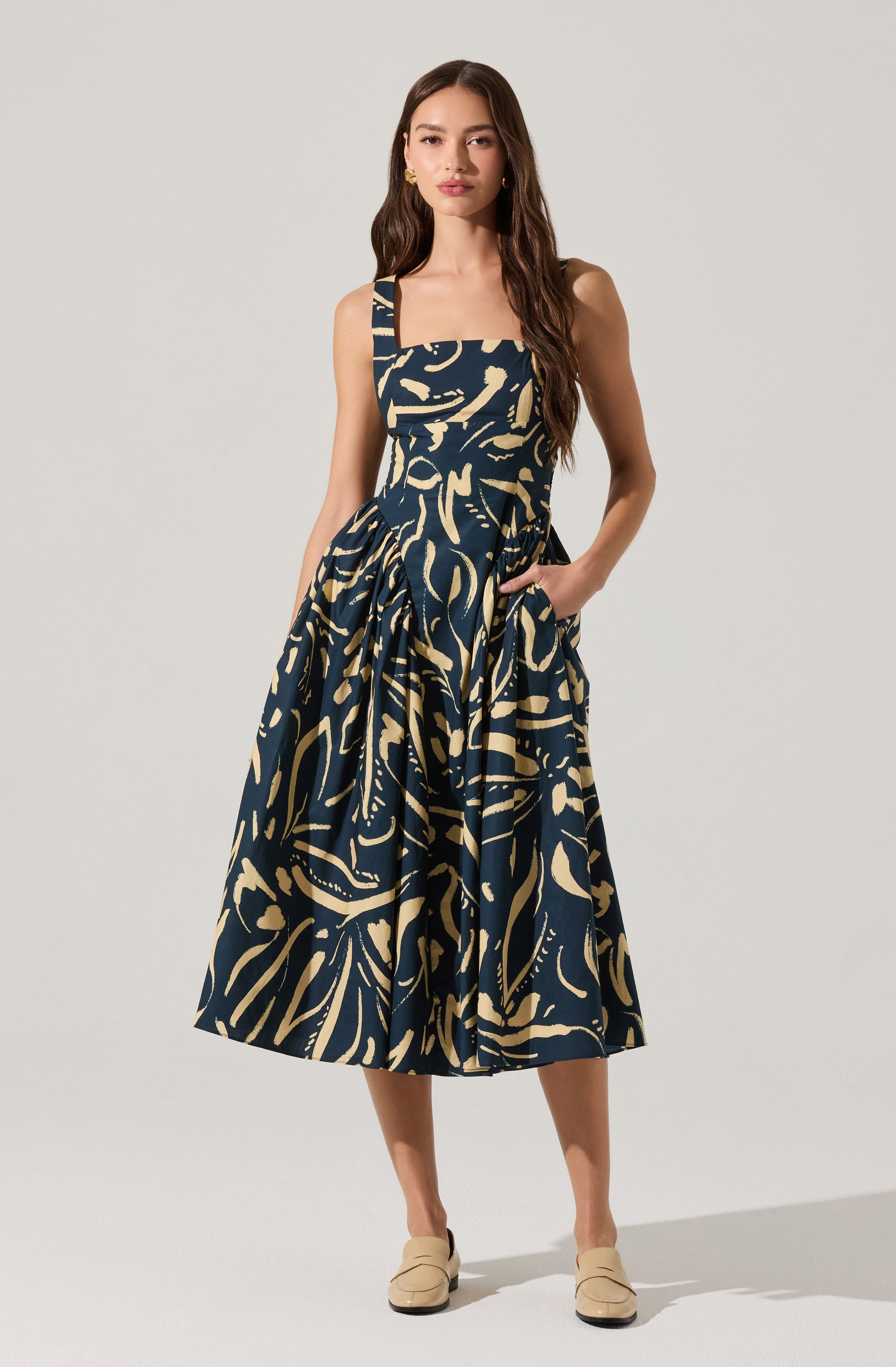 A woman is wearing a sleeveless, ankle-length dress with a square neckline and wide shoulder straps. The dress features a dark navy blue base color with an abstract beige pattern throughout. She pairs the outfit with beige slip-on shoes.