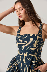 A woman is wearing a sleeveless dress with wide shoulder straps. The dress features an abstract pattern in beige and navy blue. The fitted bodice and gathered skirt give the dress a stylish and elegant look.