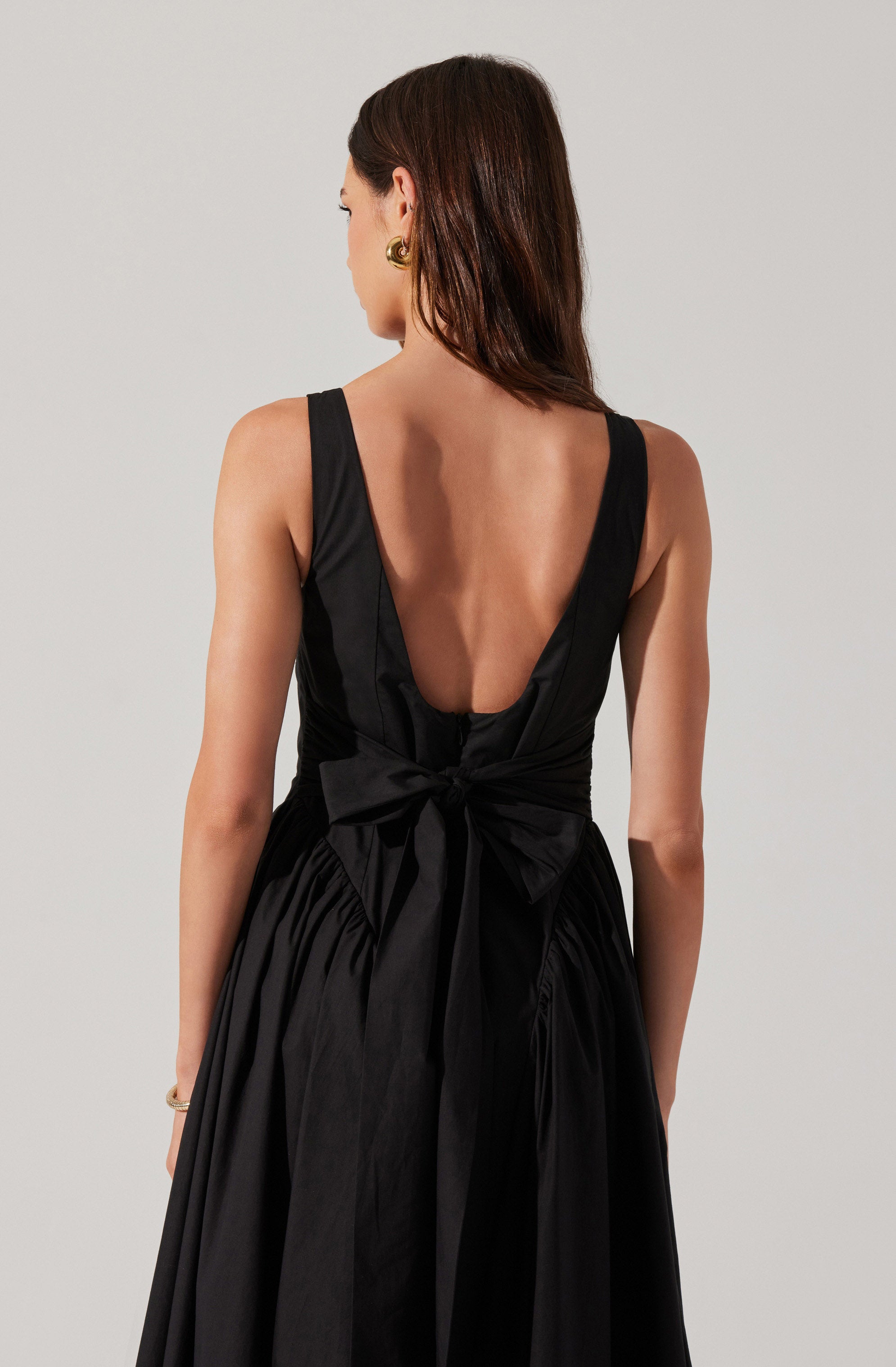 A woman is wearing a sleeveless black dress with a deep back neckline. The dress features wide shoulder straps and a large bow tied at the waist. The fabric appears to be satin or similar material, giving it a shiny, elegant look.
