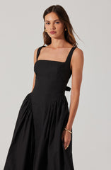 A woman is wearing a sleeveless black dress with wide shoulder straps. The dress has a fitted bodice and a gathered, full skirt. She accessorizes with gold hoop earrings, a gold bangle on her wrist, and a ring.