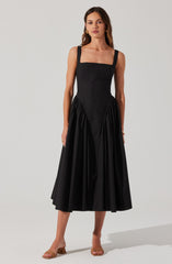 A woman is wearing a sleeveless black dress with a fitted bodice, square neckline, and wide shoulder straps. The skirt is gathered at the waist with pleats, creating a full and flowing silhouette that falls to mid-calf length. She is also wearing beige open-toe sandals with a block heel.