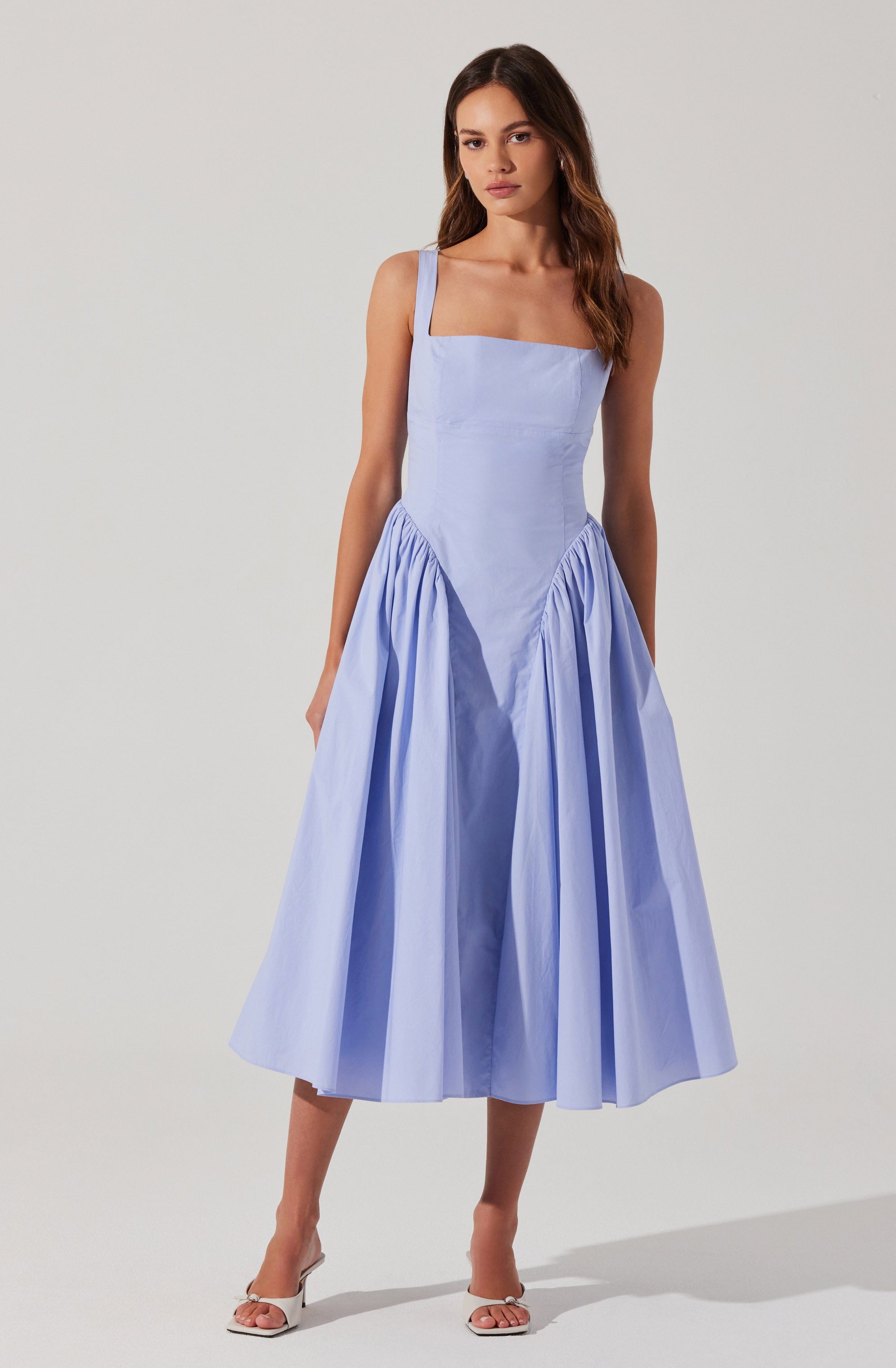 A woman is wearing a sleeveless, light blue dress with wide straps and a fitted bodice. The dress features a full, gathered skirt that falls below the knees, creating a voluminous silhouette. She is also wearing white open-toe high-heeled sandals.