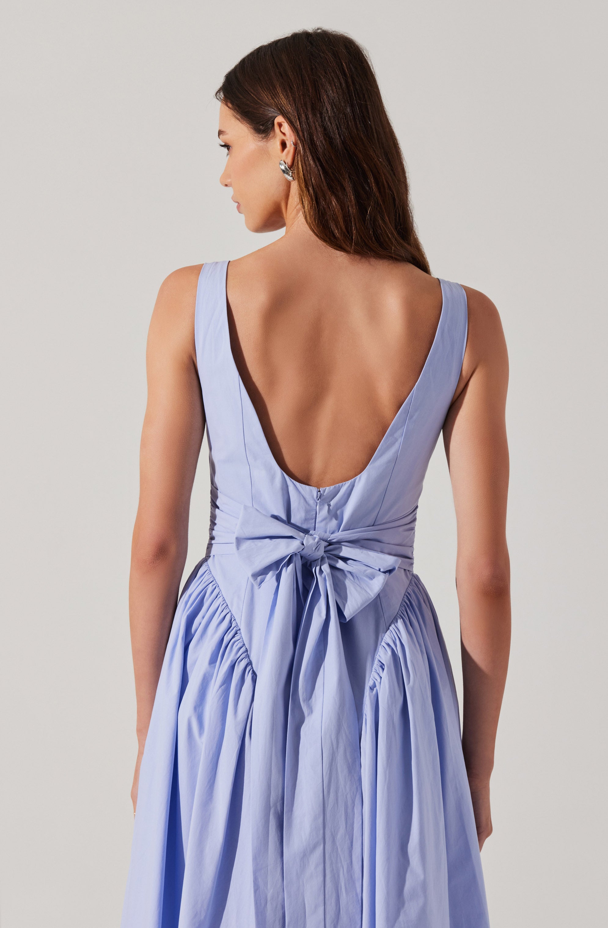 A woman is wearing a sleeveless, light blue dress with a deep V-back neckline. The dress features a fitted bodice with vertical seam details and a large bow tied at the waist. The skirt is gathered and has a full, flowing appearance with pleats.
