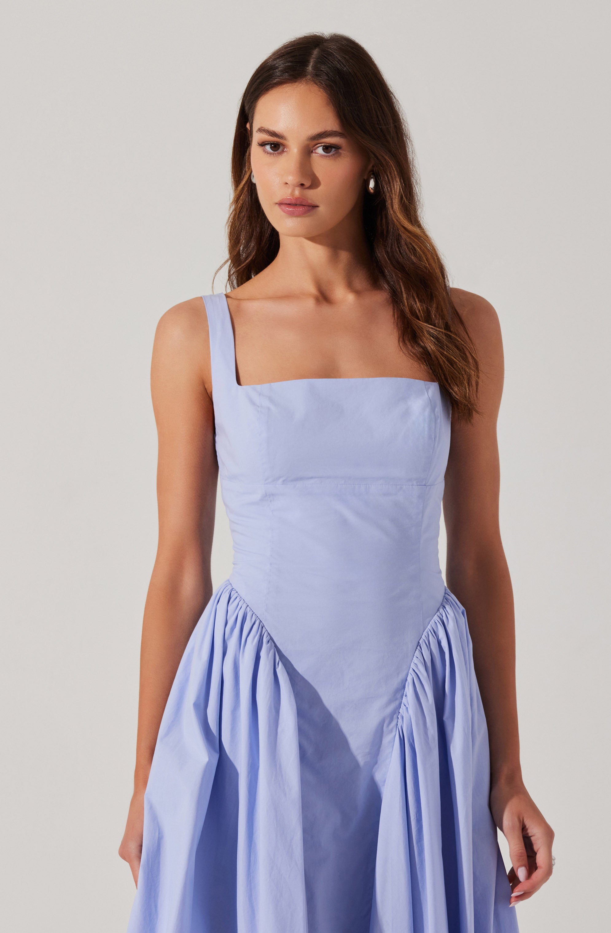 A woman is wearing a light blue sleeveless dress with wide shoulder straps and a square neckline. The fitted bodice extends to the waist, and the skirt is gathered with pleats, creating a voluminous effect. The fabric appears smooth and slightly shiny.