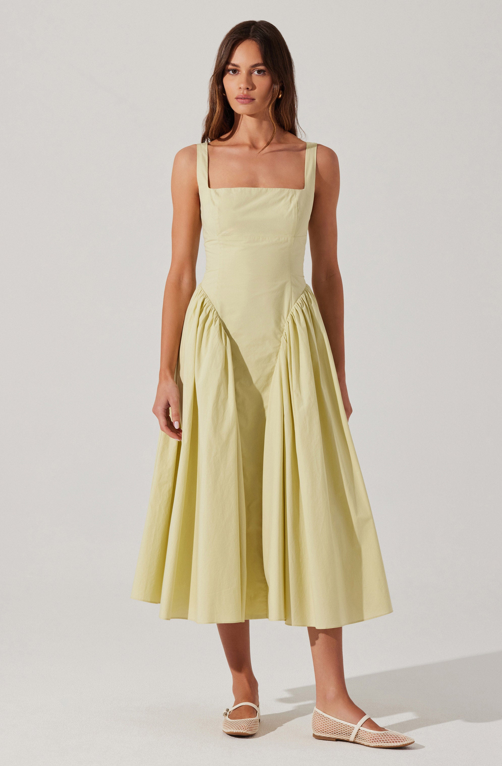 A woman is wearing a light yellow sleeveless dress with wide straps and a fitted bodice. The dress features a rectangular sheer panel across the chest and a full, gathered skirt that falls to mid-calf length. She is also wearing light-colored, open-toe, mesh flats.