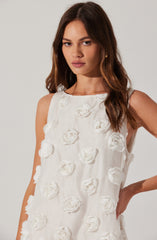 A woman wearing a sleeveless white dress decorated with 3D floral patterns that resemble roses. The dress has a high, round neckline and a relaxed fit. She has long, wavy brown hair and a neutral expression.