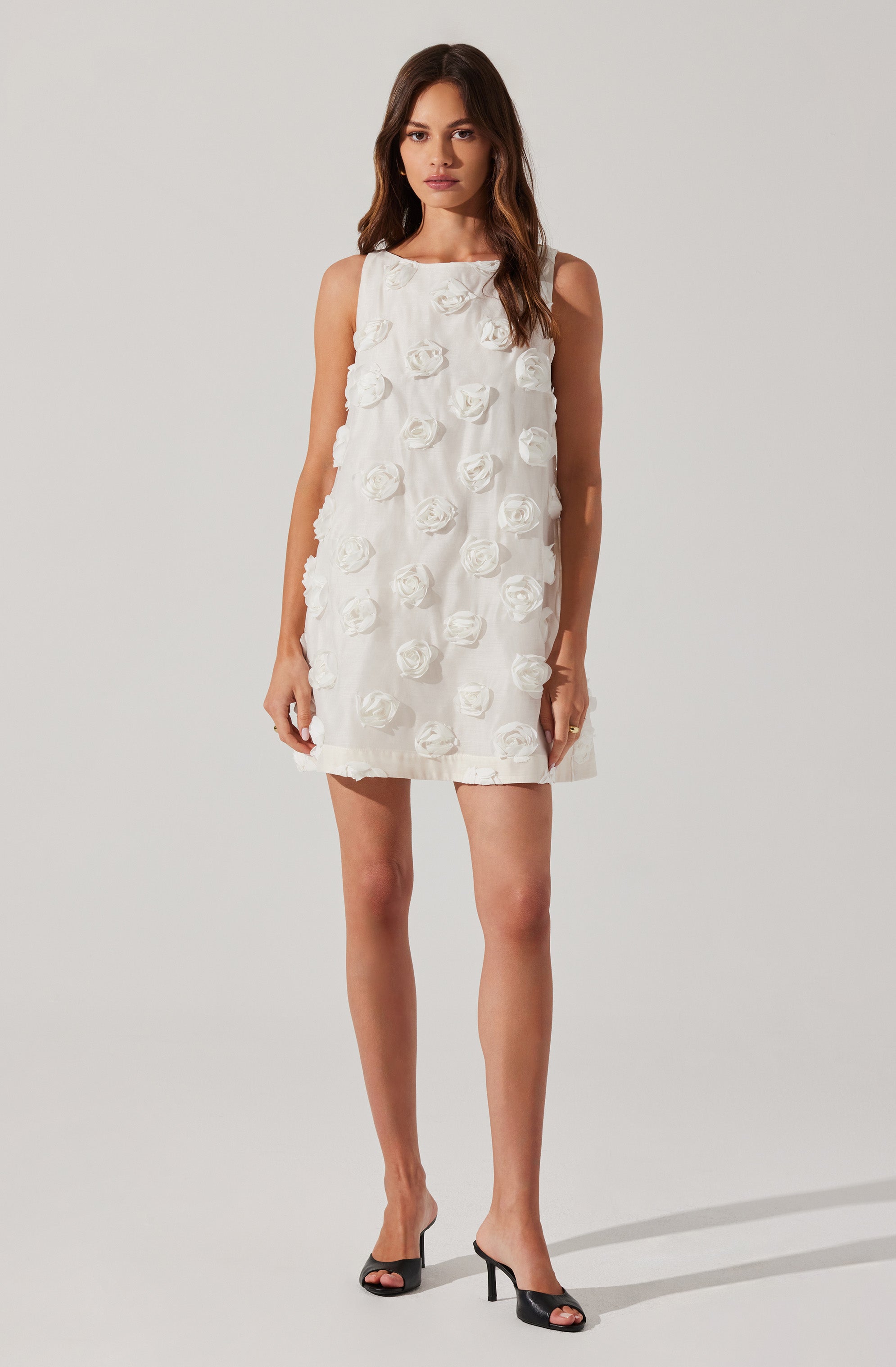 A woman is wearing a sleeveless, knee-length white dress with a round neckline. The dress features textured, three-dimensional floral appliqués all over the fabric. She is also wearing black open-toe high-heeled shoes.