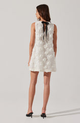The dress is sleeveless and white, decorated with textured, three-dimensional floral patterns made of fabric roses throughout. It features a black bow at the neckline, with the tie hanging down the back. The dress has a loose, A-line silhouette and ends above the knees.