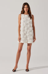 A woman is wearing a sleeveless, knee-length, white dress decorated with 3D floral appliqués. She is also wearing black open-toe high heels.