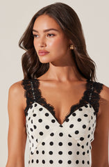 A woman is wearing a sleeveless, white dress with black polka dots. The dress features black lace trim along the V-neckline and shoulder straps.