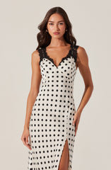 A woman is wearing a sleeveless, floor-length dress with black polka dots on a white background. The dress features black lace detailing on the shoulder straps and a V-neckline. There is a slit on the side of the skirt, revealing a part of her leg. The overall style of the dress appears elegant and quaint.