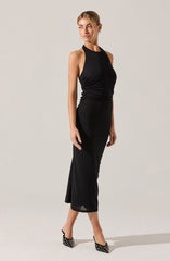 A woman is wearing a sleeveless black halter-neck dress that is fitted around the waist with ruched detailing and a long, flowing skirt that reaches mid-calf. She is also wearing black high-heeled shoes adorned with metallic eyelet details.