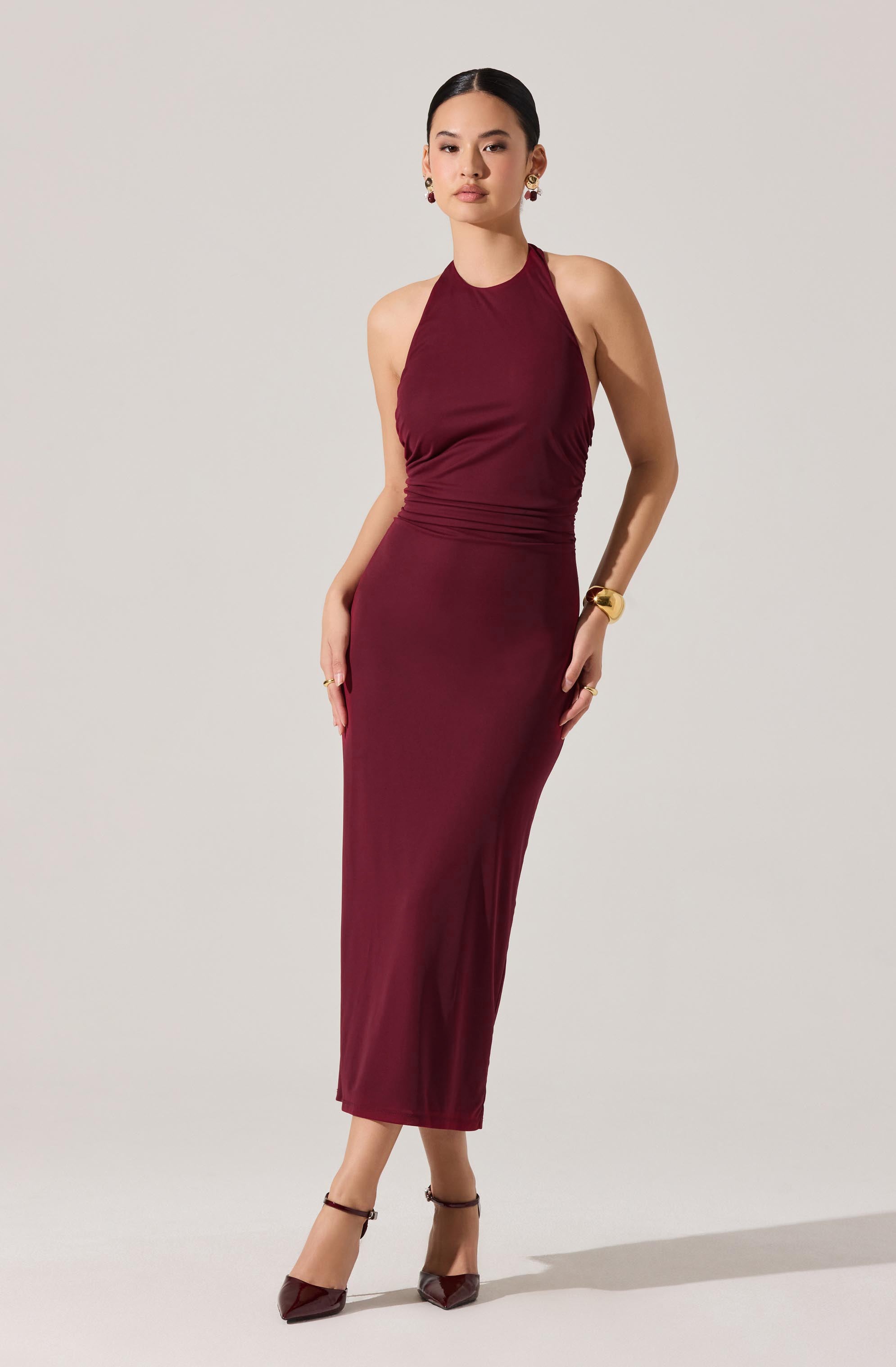 A woman is wearing a sleeveless, high-neck, ankle-length dress in a deep burgundy color. The dress is fitted at the waist with ruching details around the midsection. She is accessorized with earrings, a gold wristwatch, and rings. She is also wearing matching burgundy high heels with ankle straps.