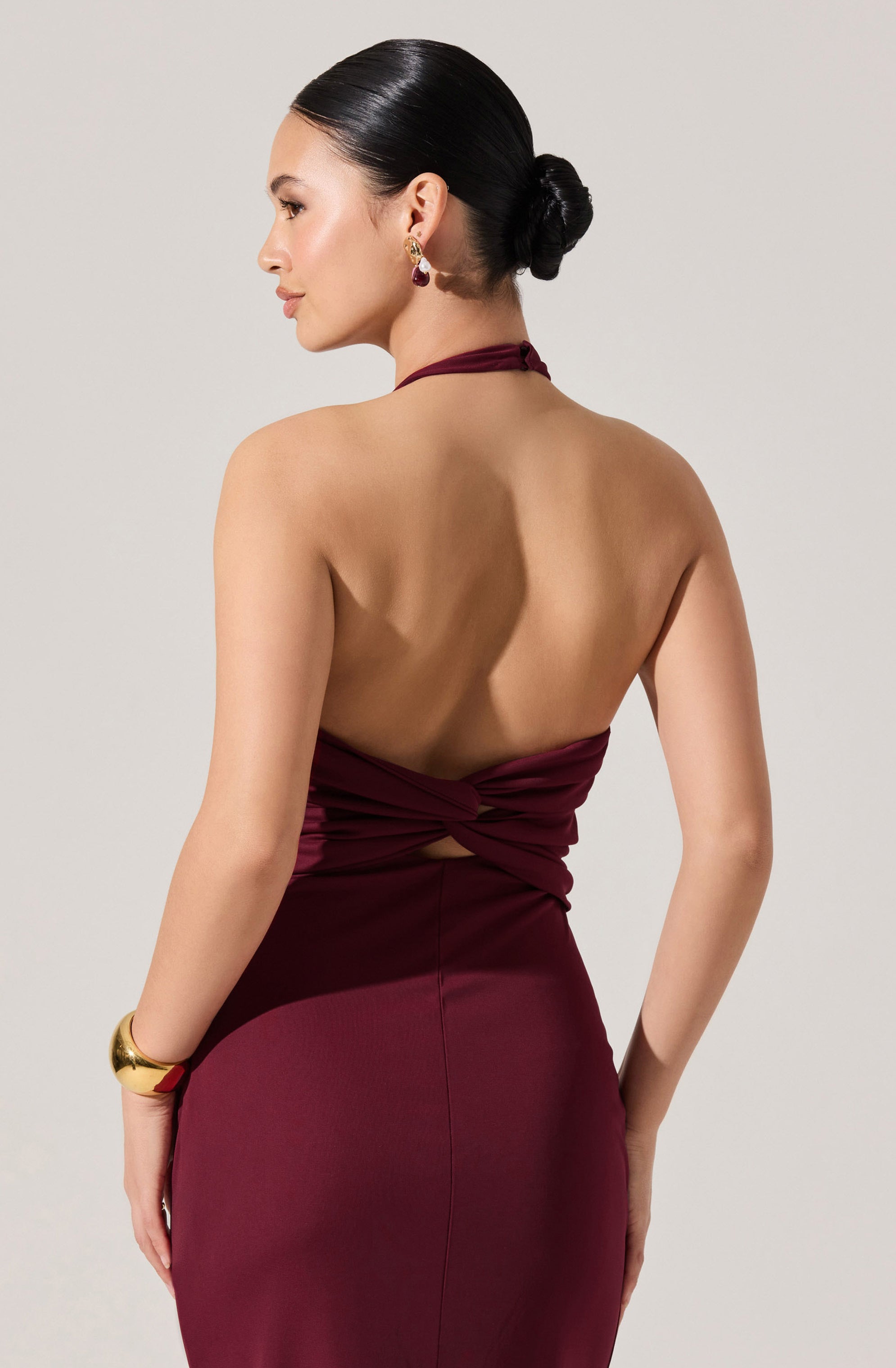 A woman is wearing a sleeveless, backless, burgundy dress with a halter neckline. The dress has a fitted silhouette and features a twist detail at the back. She has her hair styled in a sleek, low bun and is accessorizing with gold earrings and a wide gold bracelet on her left wrist.
