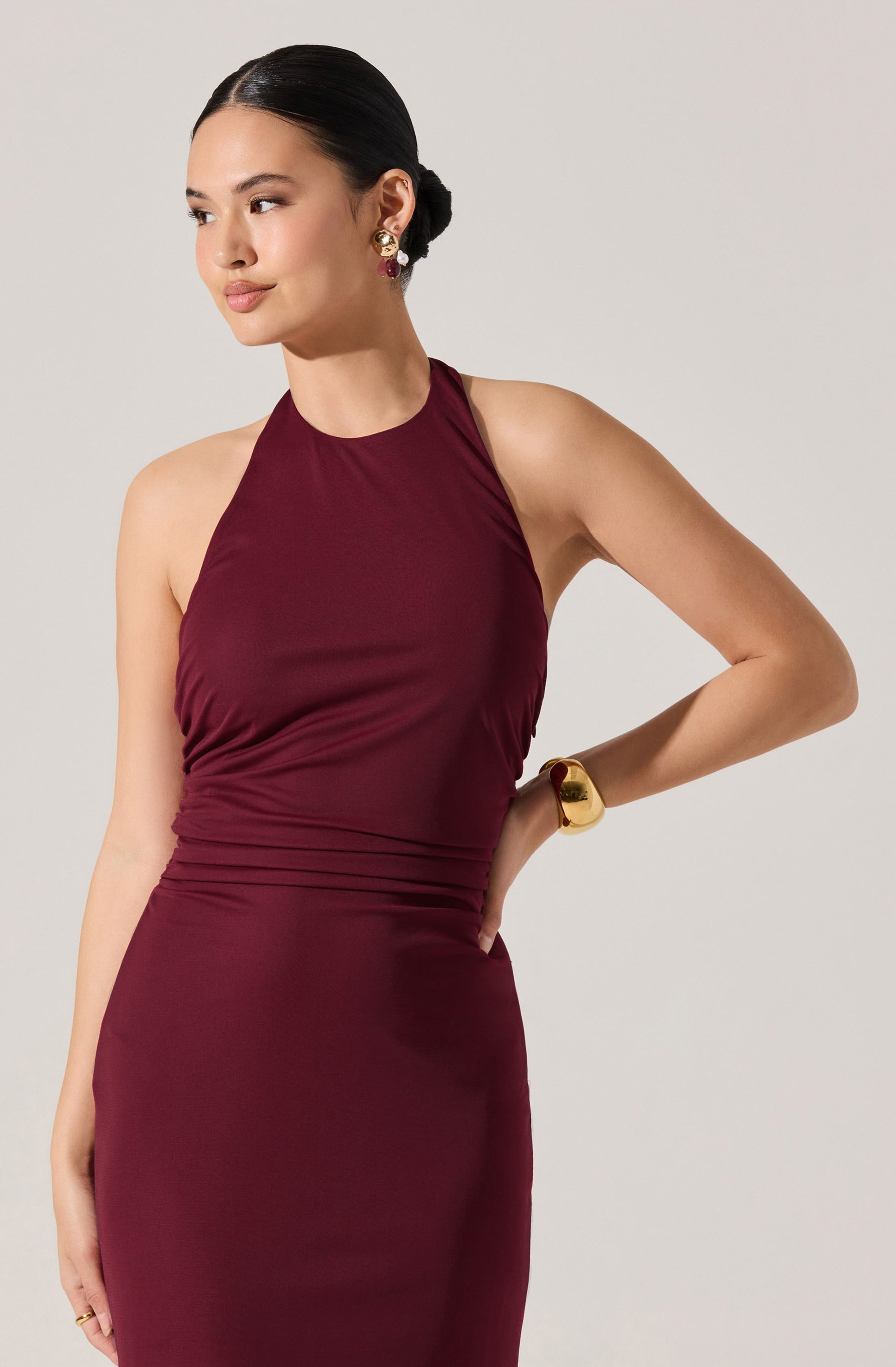 A woman is wearing a sleeveless, fitted dress in a deep burgundy color. The dress has a high neckline and is made of a smooth, slightly shiny fabric. It features ruching details around the waist and bust area.