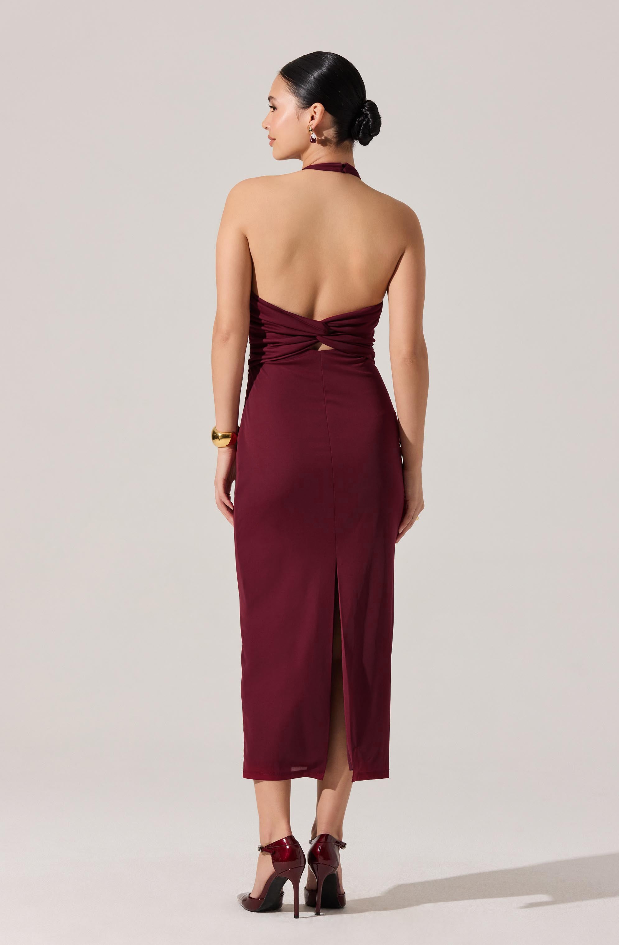 A woman is wearing a sleeveless, backless, burgundy dress that has a ruched bodice with a cutout detail at the waist and a slit at the back. She also has on matching burgundy high-heeled shoes and gold jewelry.