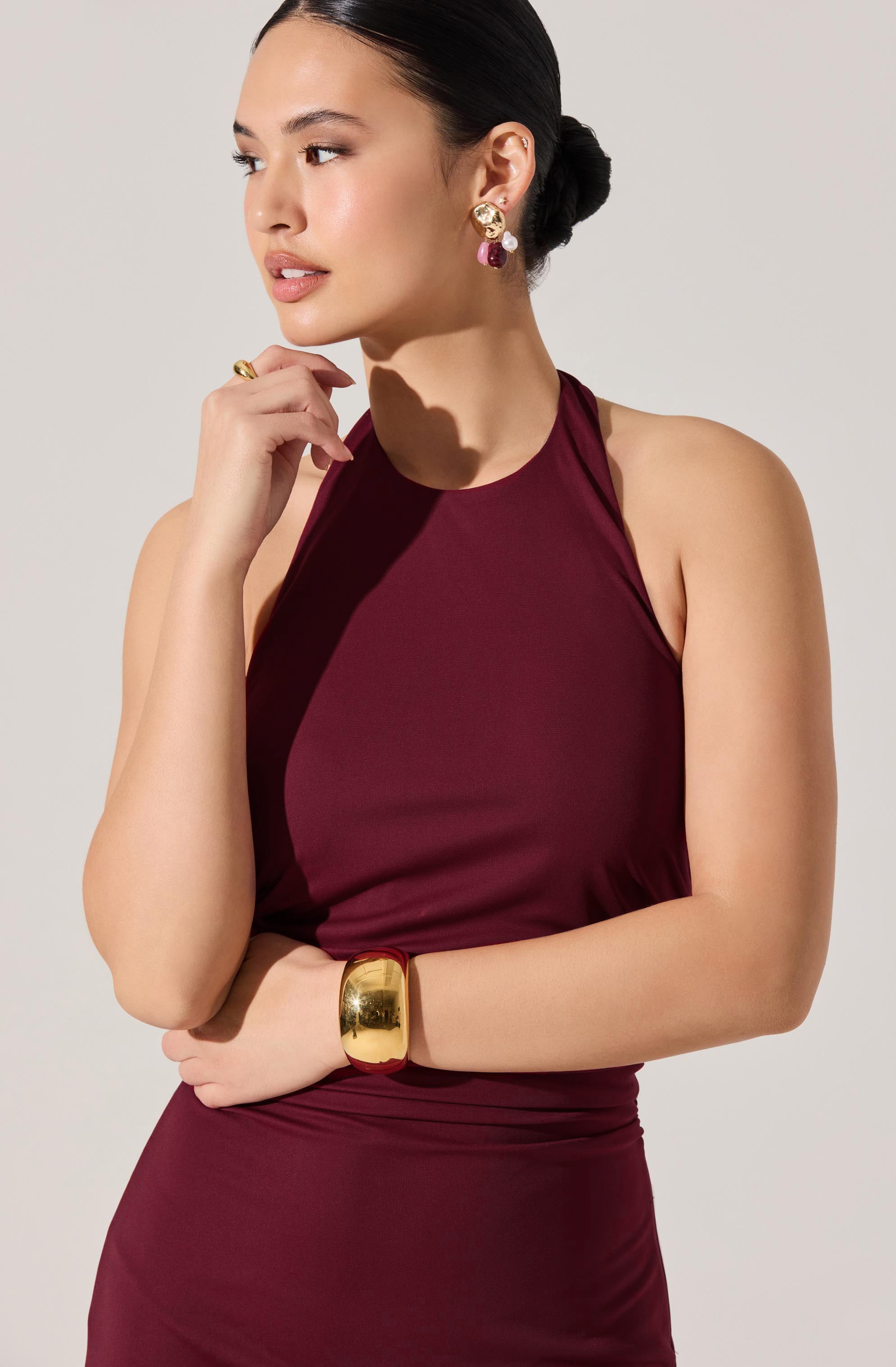 A woman is wearing a sleeveless, high-neck burgundy dress. She has layered earrings featuring gold, purple, and pink elements, a wide gold bracelet on her right wrist, and a ring on her left hand. Her hair is pulled back into a neat bun. She has a thoughtful expression with her left hand gently touching her chin.