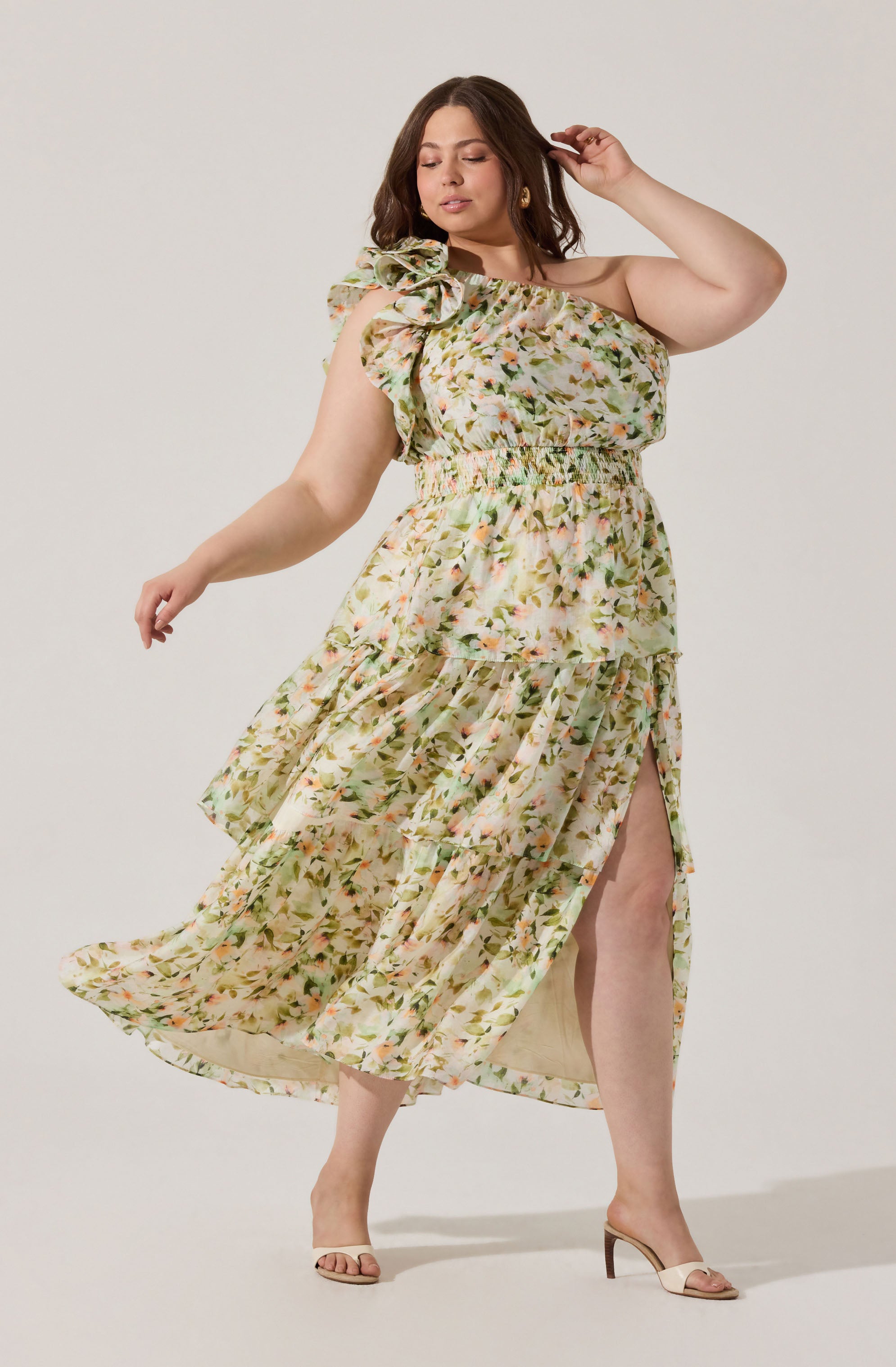 A woman is wearing a light, flowy, floral dress with a cream-colored background and pink, green, and orange flower patterns. The dress features a one-shoulder design with a large ruffle detail on the shoulder, an elastic waistband, and a high slit on the side revealing her leg. She is also wearing nude-colored open-toe high heels.